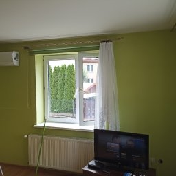 Piotr Bigos - Pokój z zielonymi ścianami po malowaniu, widoczne okno z firanką, drabina malarska i telewizor. Świeżo pomalowane ściany i czyste okno.