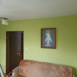 Piotr Bigos - Jasnozielony pokój z brązowymi drzwiami i obrazem w ramie nad kanapą. Widoczna lampa sufitowa i fragment drabiny malarskiej. Ściana świeżo pomalowana.