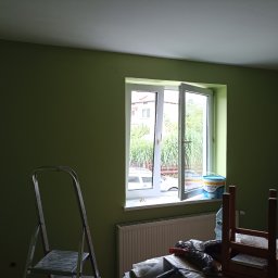 Piotr Bigos - Ściana pomalowana na zielono, widoczne otwarte okno, drabina malarska i akcesoria malarskie. Świeżo pomalowane wnętrze z widocznymi narzędziami pracy.