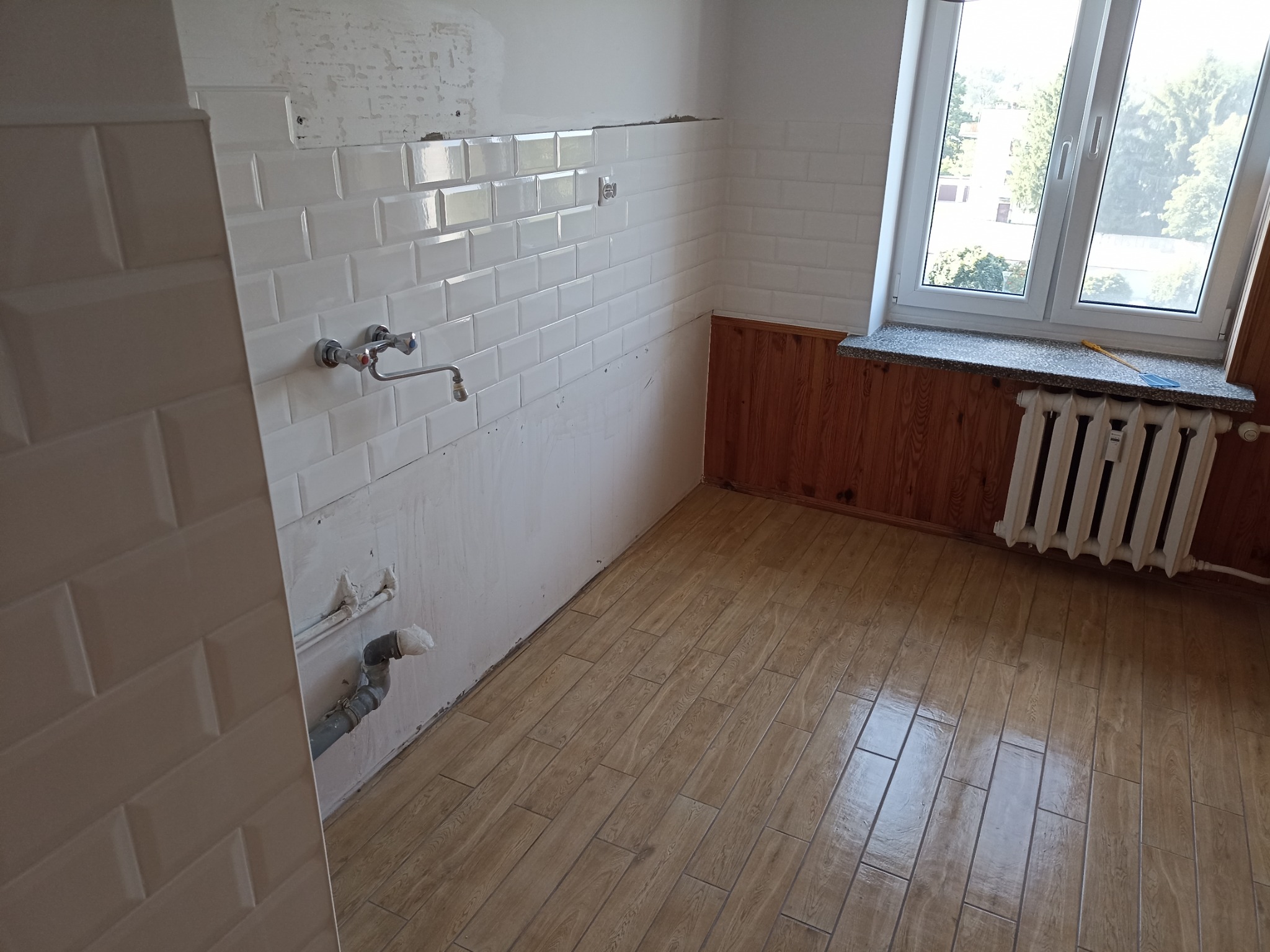 Kuchnia w trakcie remontu: białe, prostokątne płytki na ścianie do połowy wysokości, drewnopodobna podłoga, okno z widokiem na zieleń. Widoczne rury i niedokończone ściany.