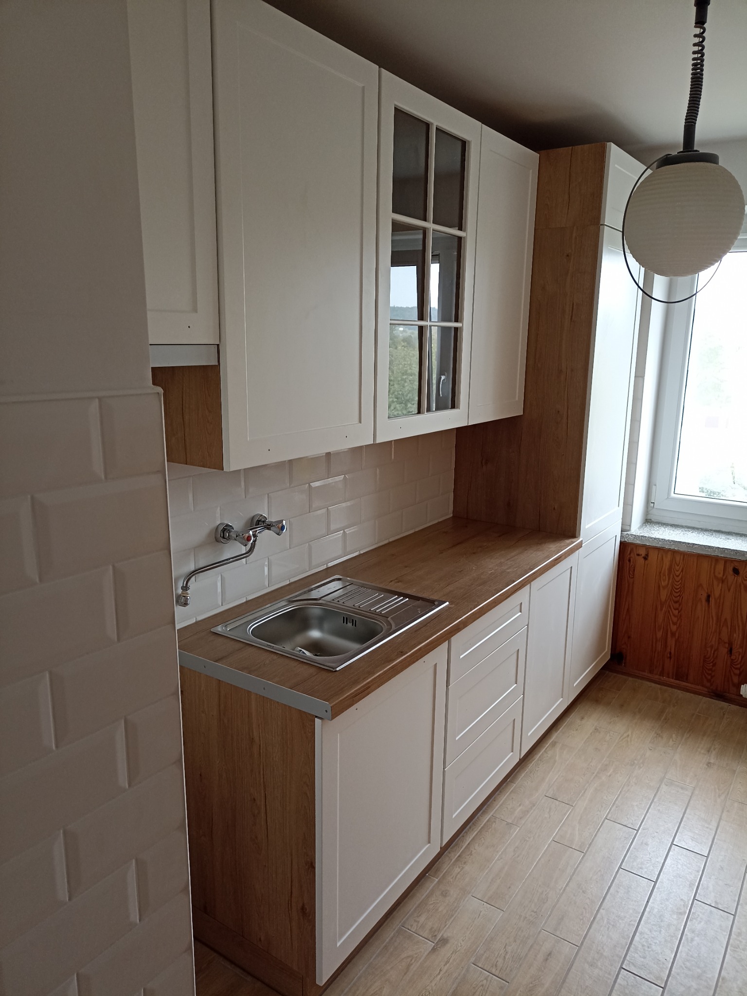 Jasna kuchnia po remoncie: białe szafki z drewnianymi akcentami, stalowy zlew, beżowe płytki na ścianie i podłodze. Widoczne okno i lampa.