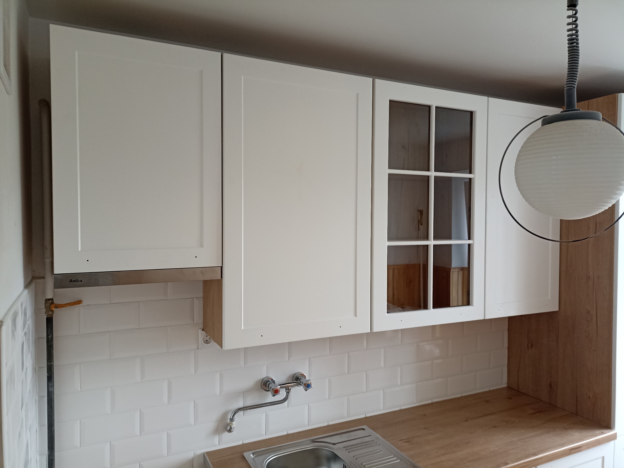 Białe, wiszące szafki kuchenne z przeszklonym frontem, okap Amica, białe płytki typu metro i drewniany blat. Ujęcie na fragment kuchni po montażu.