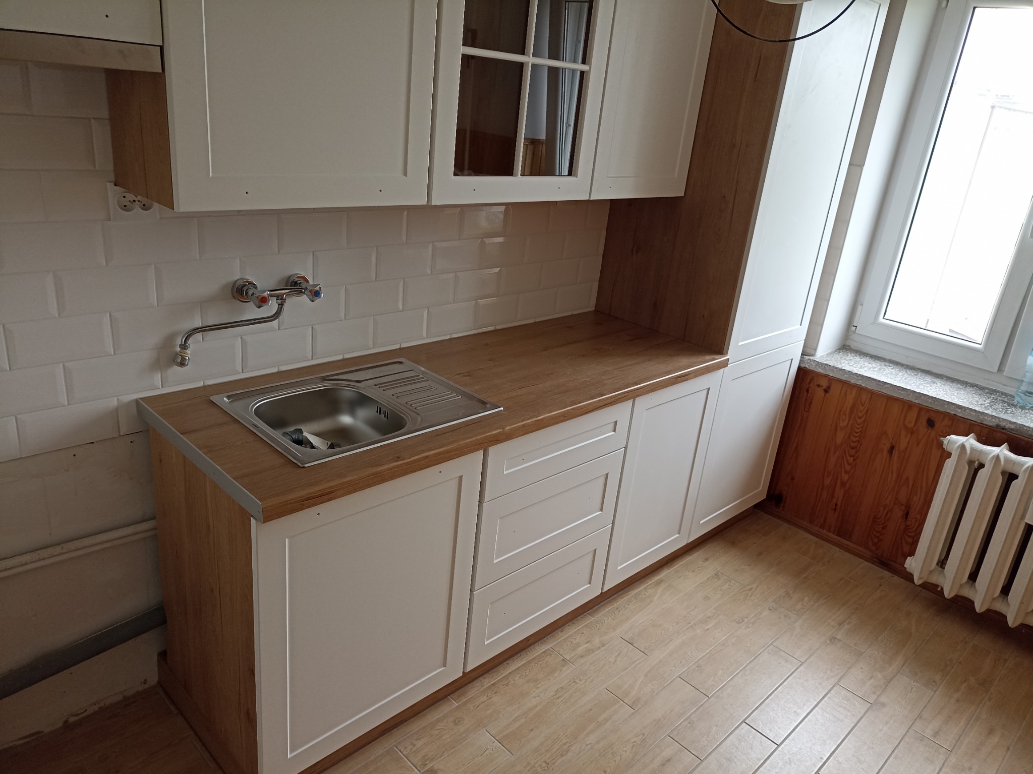 Jasna kuchnia z drewnianym blatem i białymi szafkami, widok na zlew i baterię. W tle białe płytki na ścianie i okno z grzejnikiem.