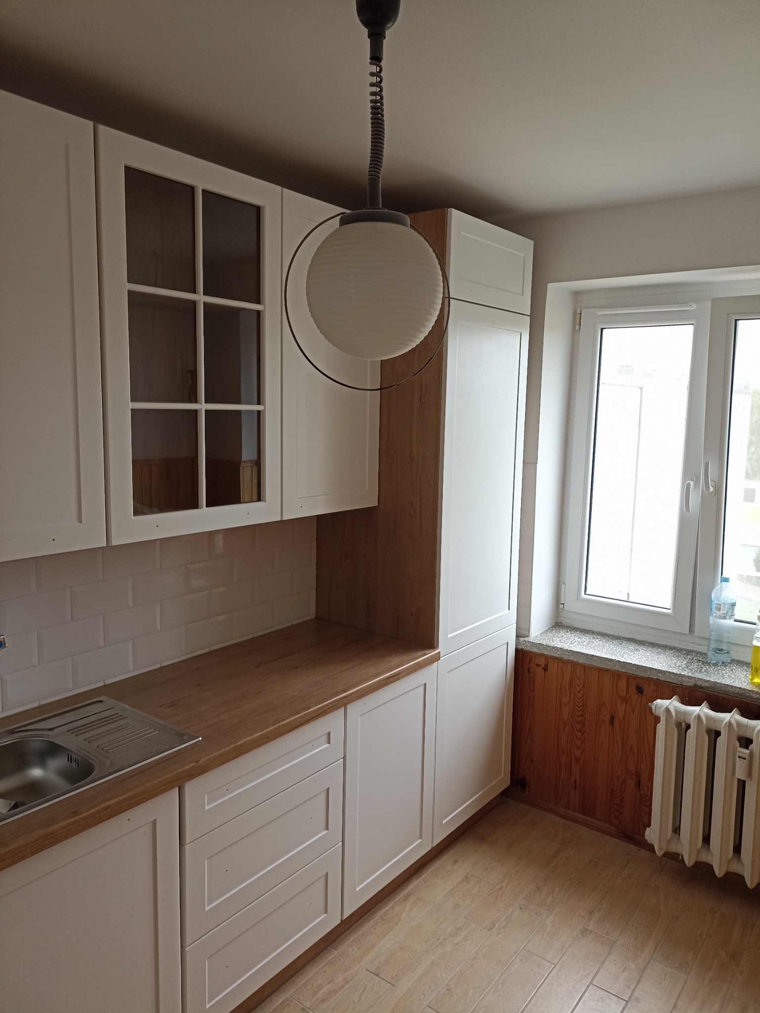 Jasna kuchnia po remoncie: białe szafki z drewnianym blatem, białe płytki na ścianie, drewniana boazeria pod oknem i grzejnik. Nowoczesne oświetlenie.