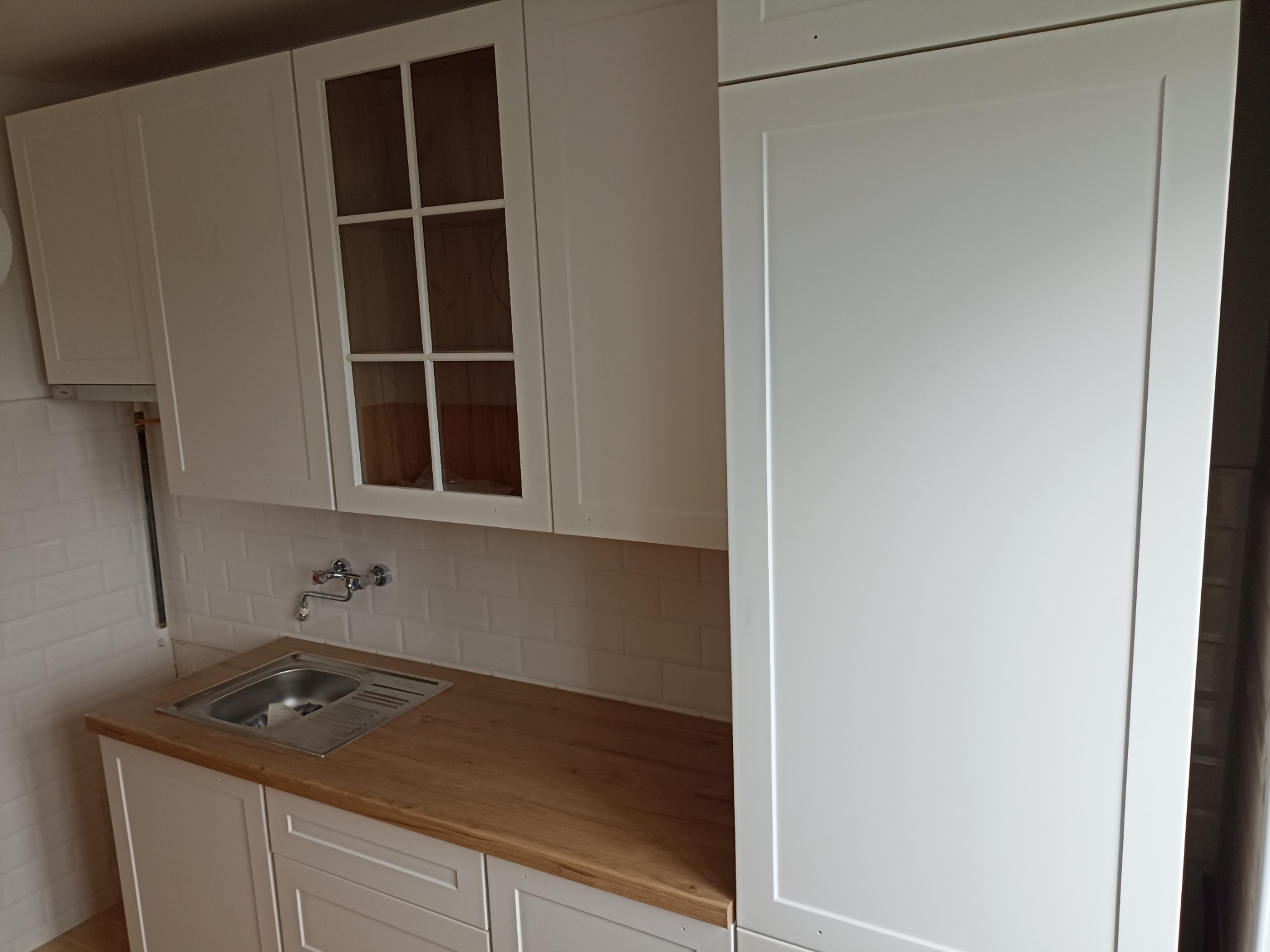 Biała zabudowa kuchenna z drewnianym blatem i chromowaną baterią, widoczna na tle białych płytek. Szafka z przeszklonymi drzwiczkami. Minimalistyczny design.