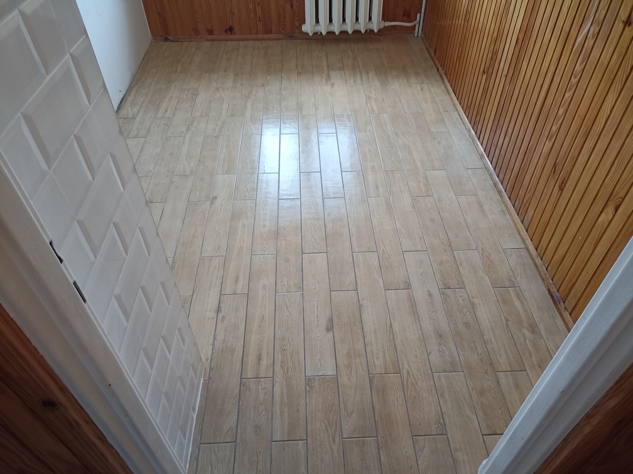 Ukończona podłoga z płytek imitujących drewno w wąskim pomieszczeniu z boazerią. Widok z góry, z fragmentem białych drzwi po lewej stronie kadru.