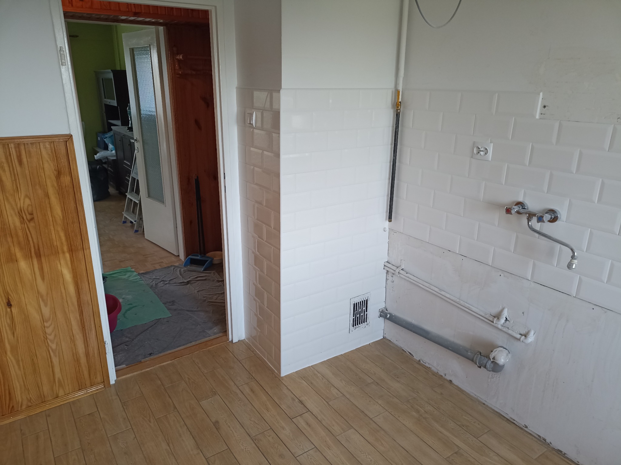 Białe, prostokątne płytki na ścianie w trakcie montażu, widoczne rury i instalacje. Podłoga wyłożona panelami imitującymi drewno. Widok na sąsiednie pomieszczenie.