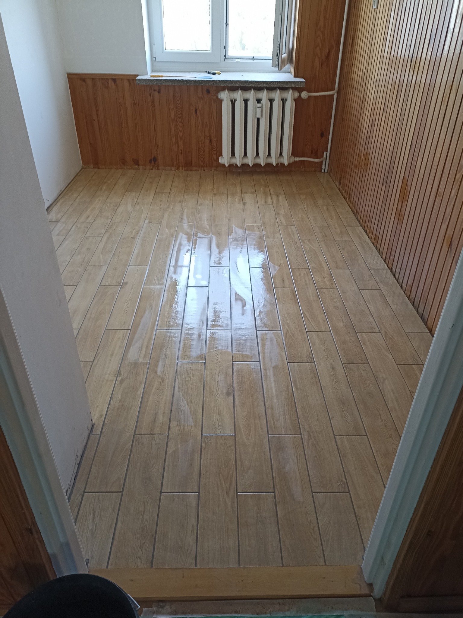 Ukończona podłoga z płytek imitujących drewno w pokoju z boazerią. Widoczny grzejnik pod oknem. Podłoga świeżo umyta, z refleksami światła.