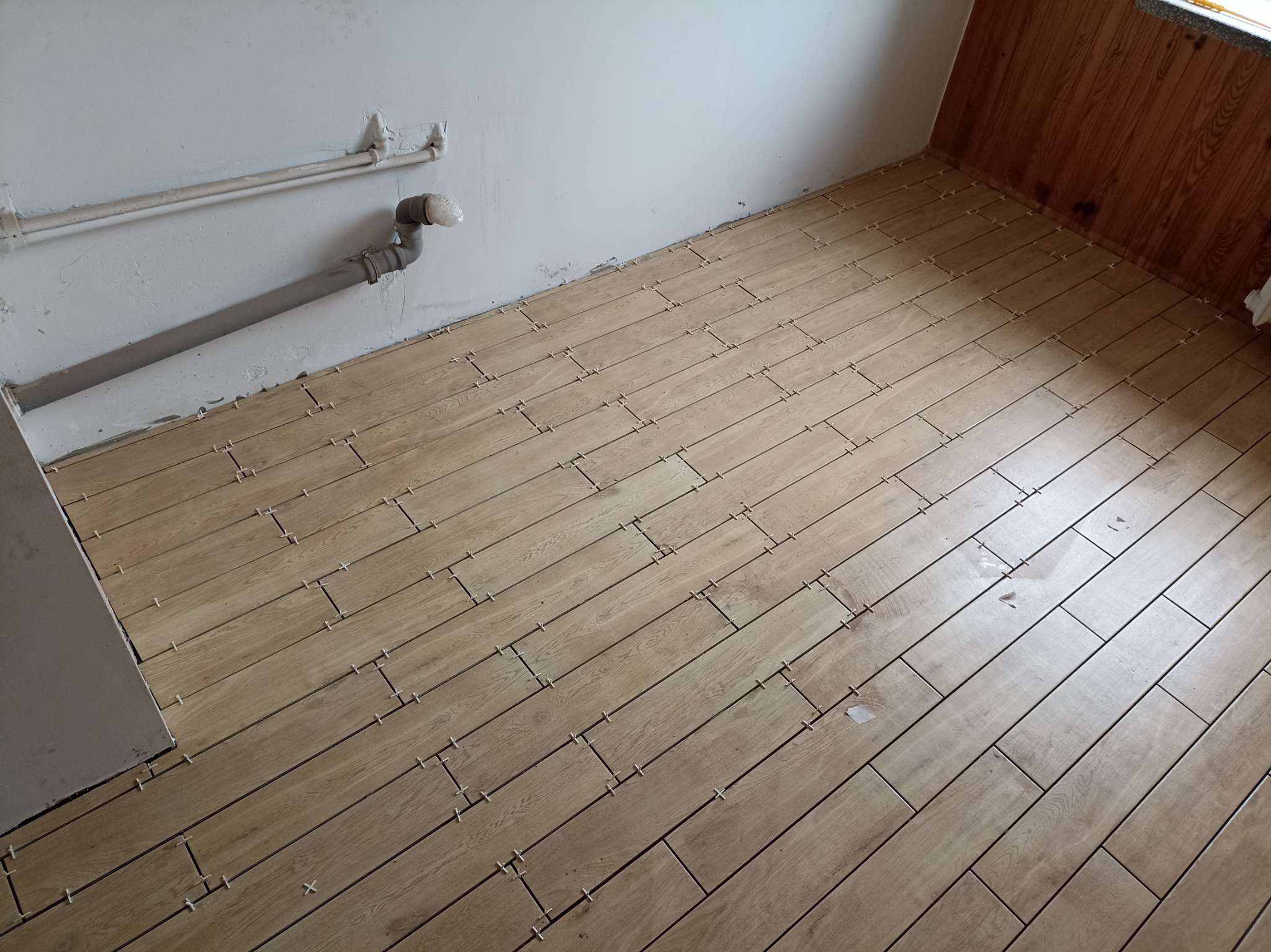 Układanie podłogi z płytek imitujących drewno, z krzyżykami dystansowymi, widoczne rury przy ścianie. Ukończony etap prac glazurniczych w pomieszczeniu.