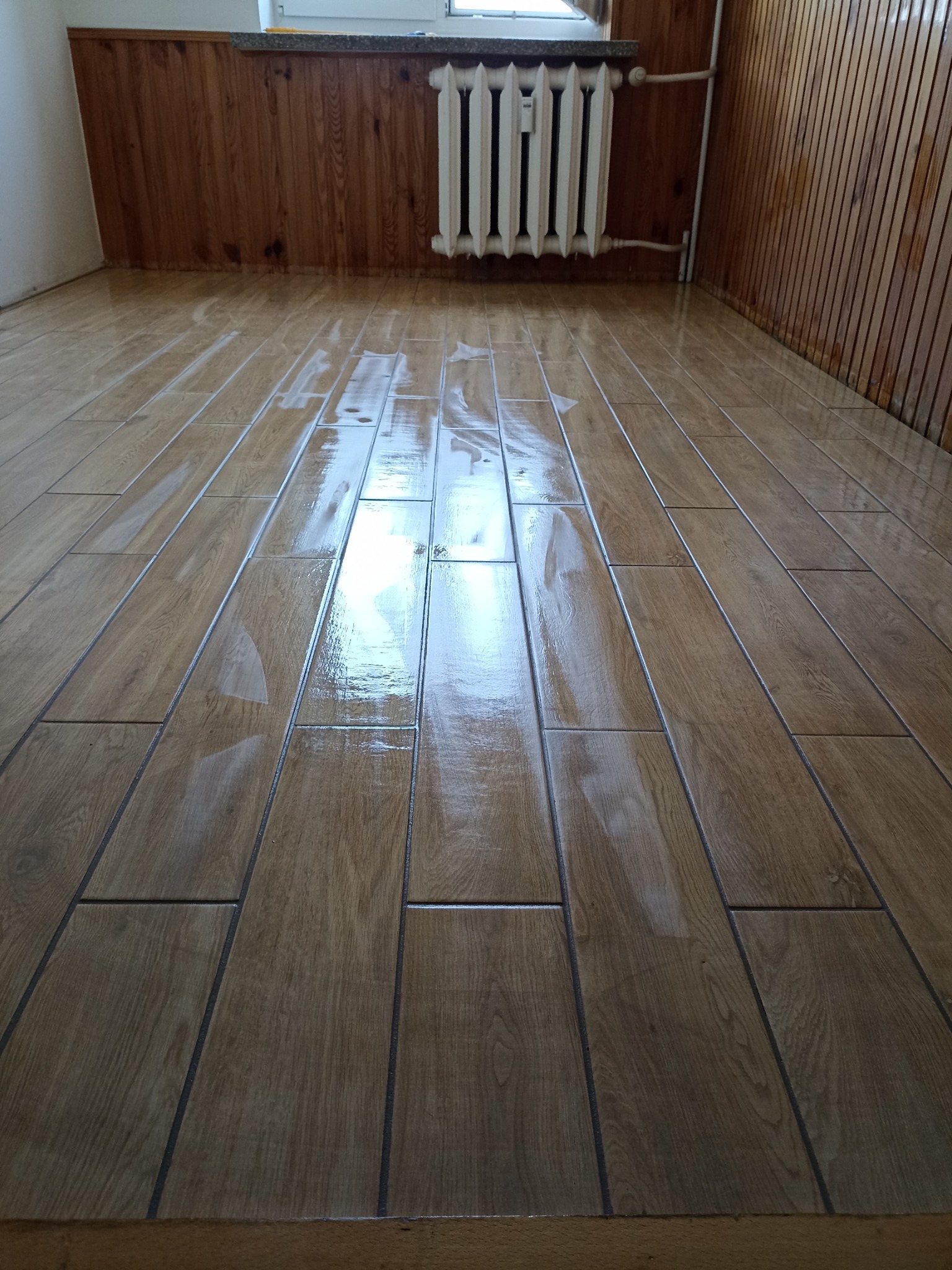 Połyskująca, drewnopodobna podłoga z płytek ceramicznych w pokoju z boazerią i grzejnikiem. Świeżo ułożona glazura, widoczne odbicia światła.