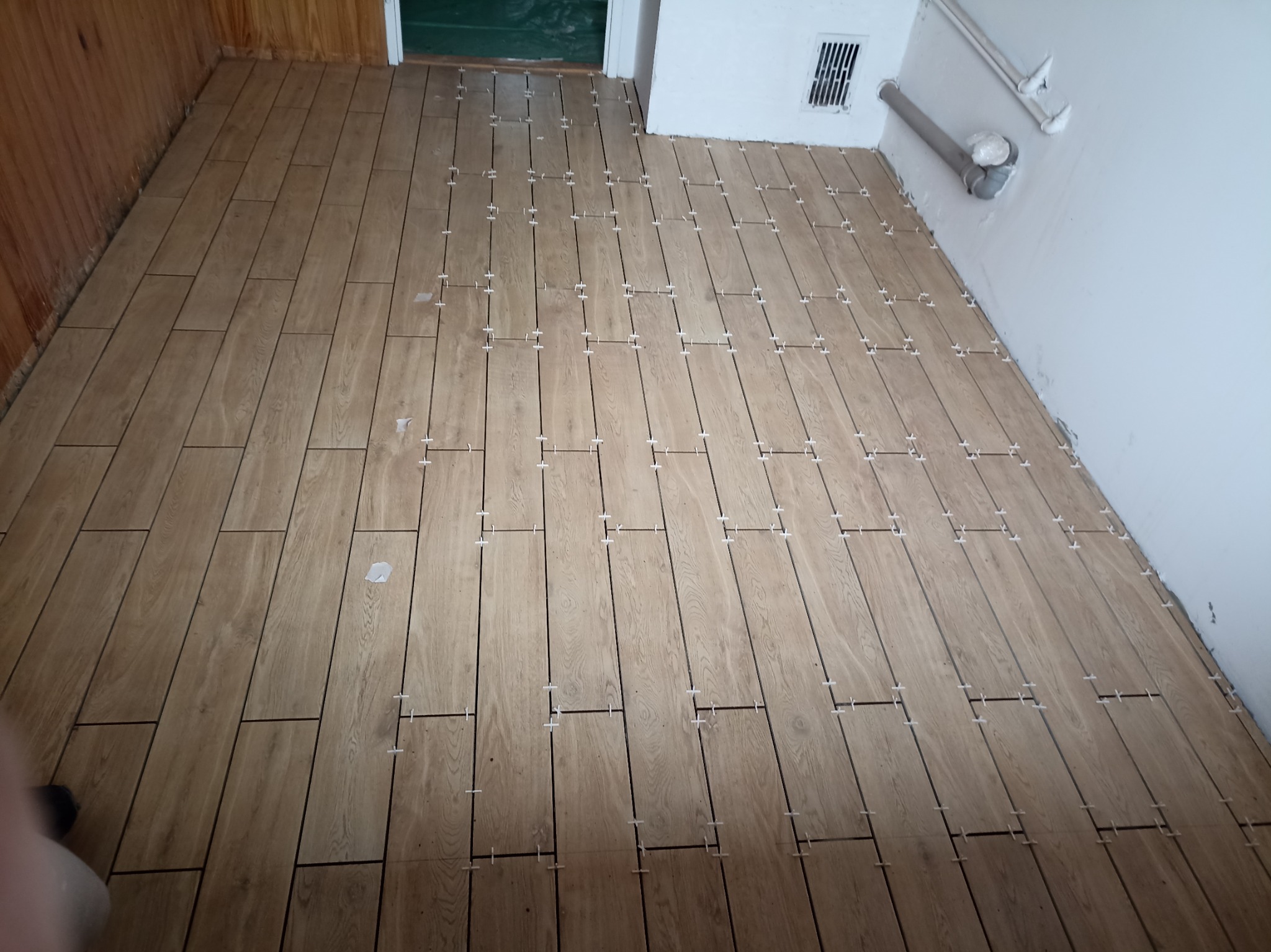 Układanie podłogi z płytek imitujących drewno, z użyciem krzyżyków dystansowych. Widoczne fragmenty ściany i rury. Jasne, naturalne oświetlenie.