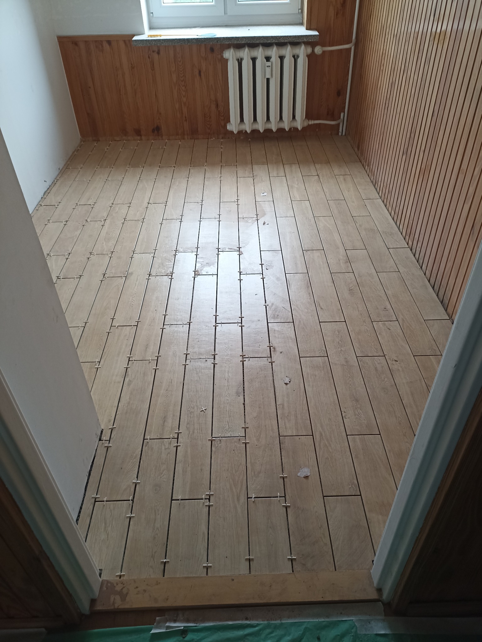 Układanie podłogi z płytek imitujących drewno w pomieszczeniu z boazerią. Widoczne krzyżyki dystansowe. Ujęcie z progu drzwi.