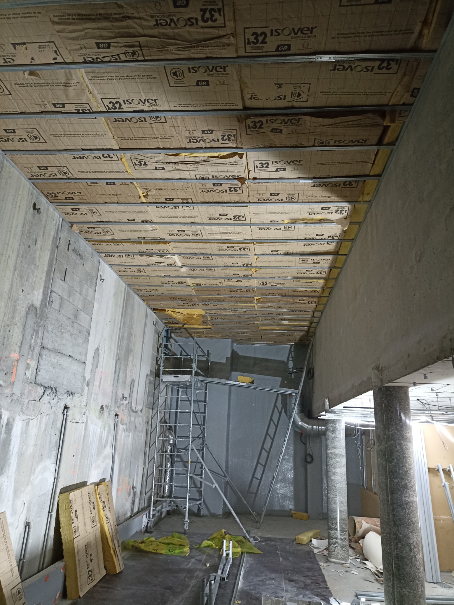 Montaż izolacji termicznej Isover na suficie podwieszanym w surowym wnętrzu. Widoczne rusztowania i materiały budowlane. Surowy beton i instalacje.