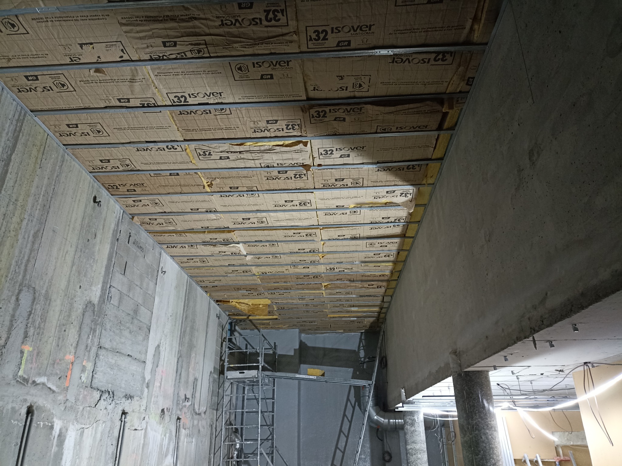 Montaż izolacji termicznej Isover 32 na metalowym ruszcie sufitowym w surowym wnętrzu. Widoczny fragment betonowej ściany i rusztowanie.
