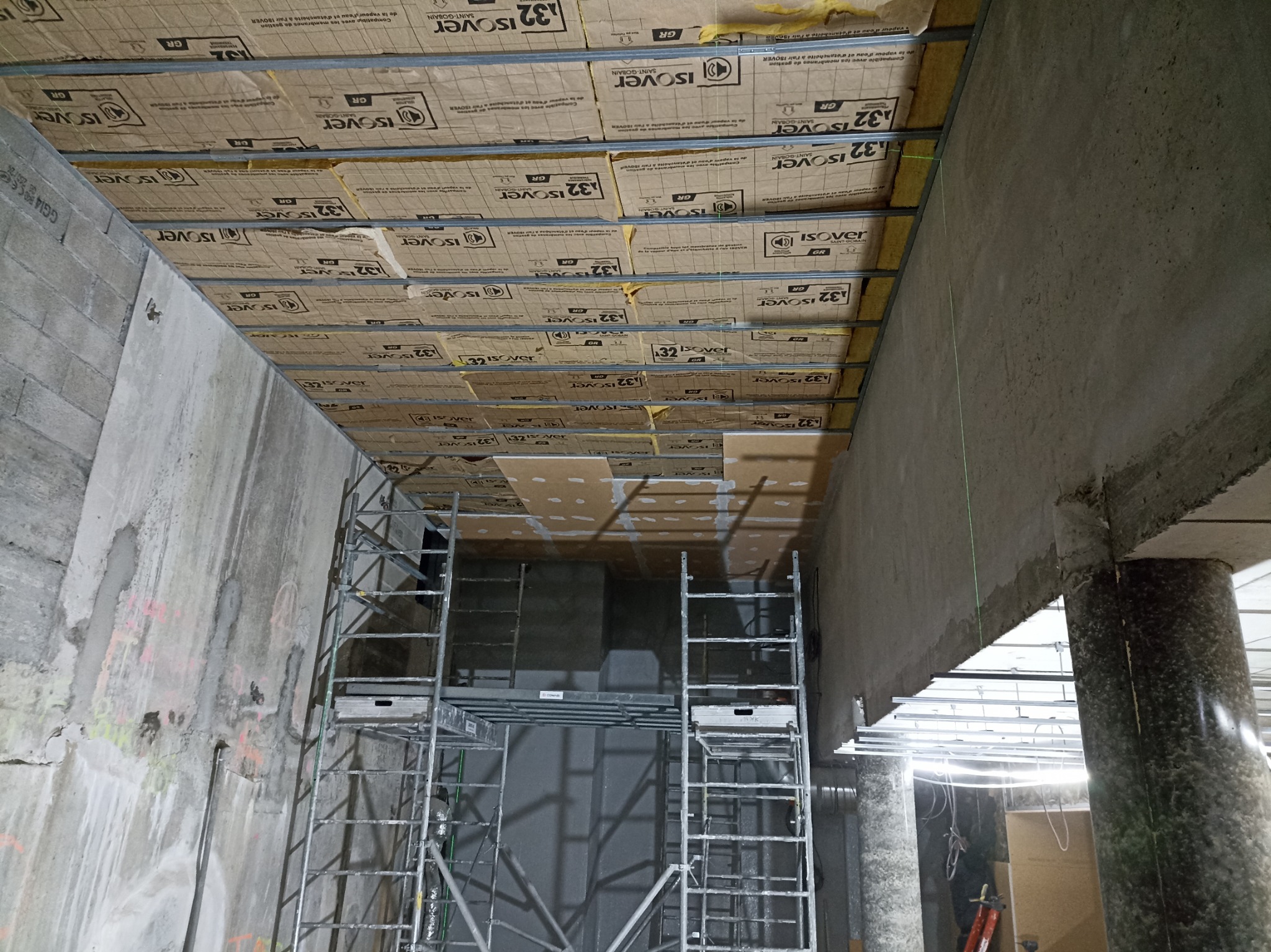 Montaż izolacji Isover 32 na metalowym ruszcie sufitowym w surowym wnętrzu. Widoczne rusztowanie i betonowe ściany. Prace wykończeniowe w toku.