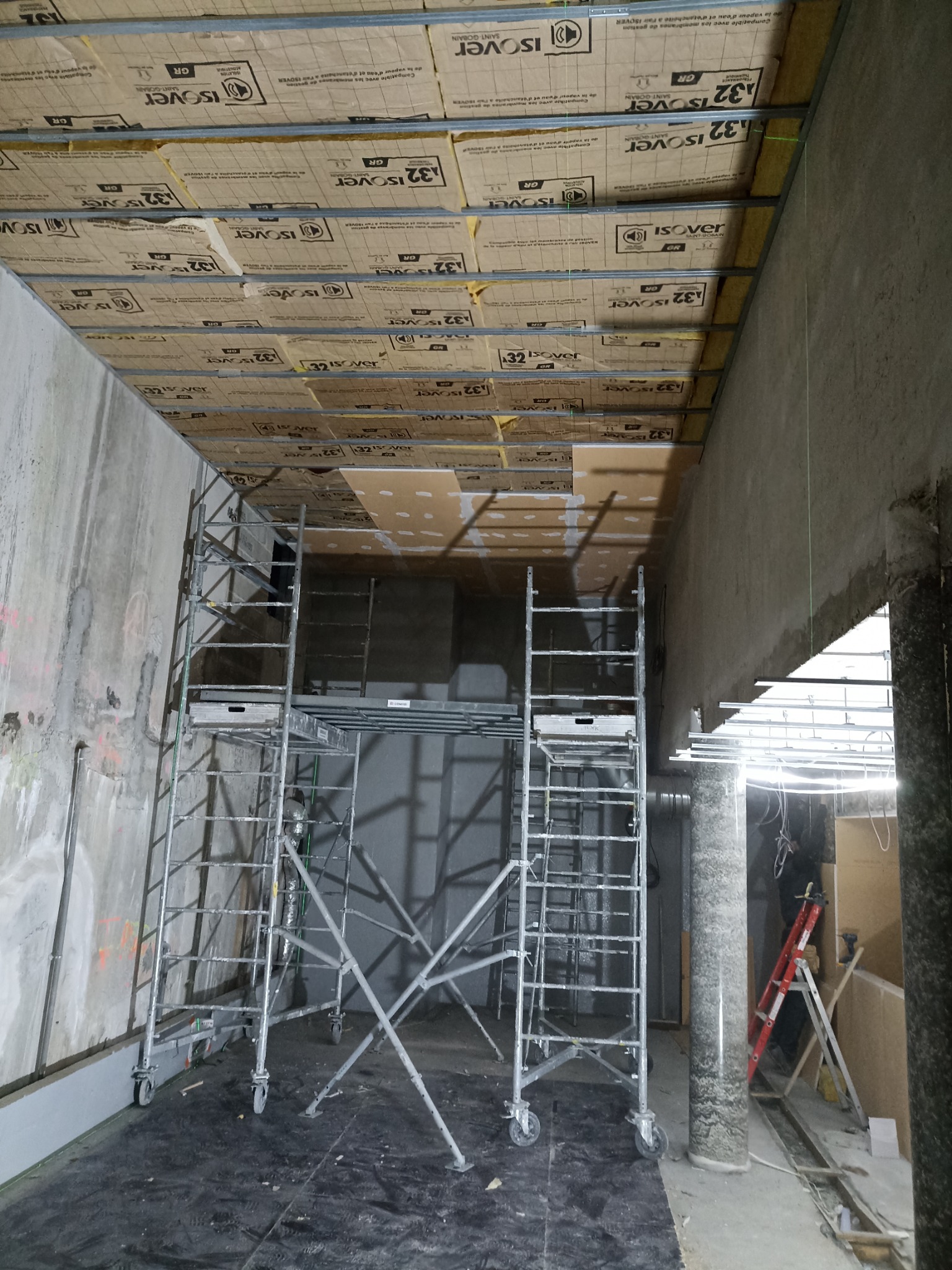 Montaż izolacji stropu z użyciem rusztowania. Widoczne płyty izolacyjne Isover i metalowa konstrukcja podwieszana. Surowe ściany w tle. Prace wykończeniowe w toku.