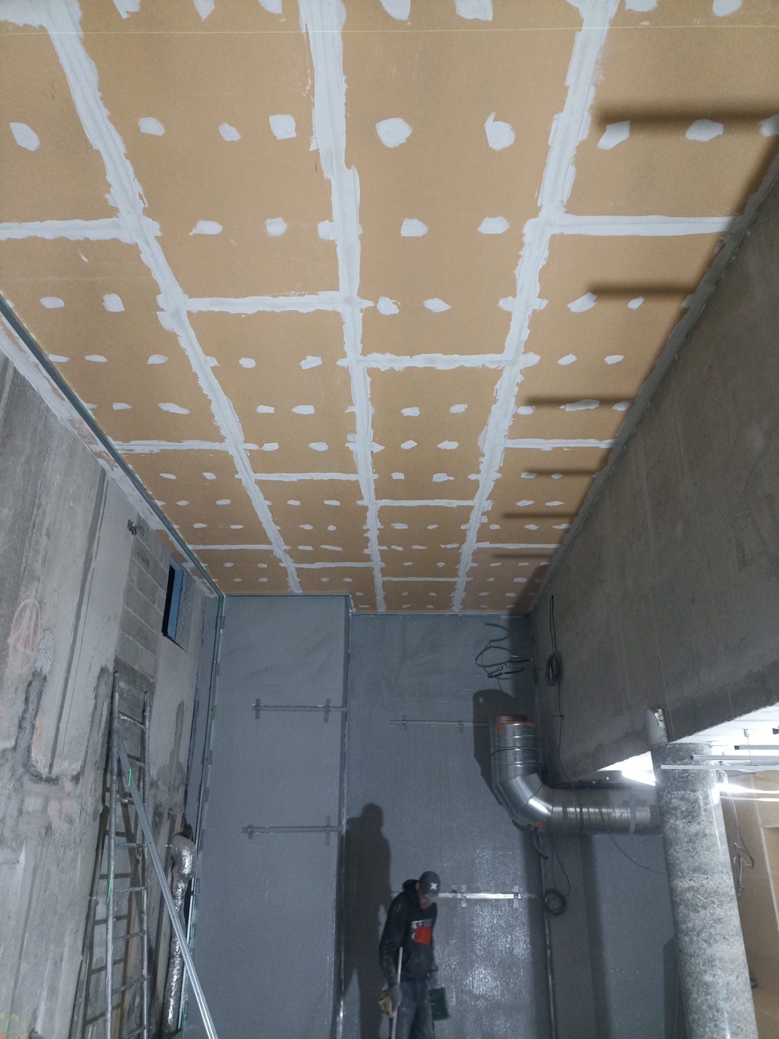 Sufit z płyt karton-gips, szpachlowane łączenia, widoczne instalacje wentylacyjne. Prace wykończeniowe w surowym wnętrzu, drabina i pracownik w tle.