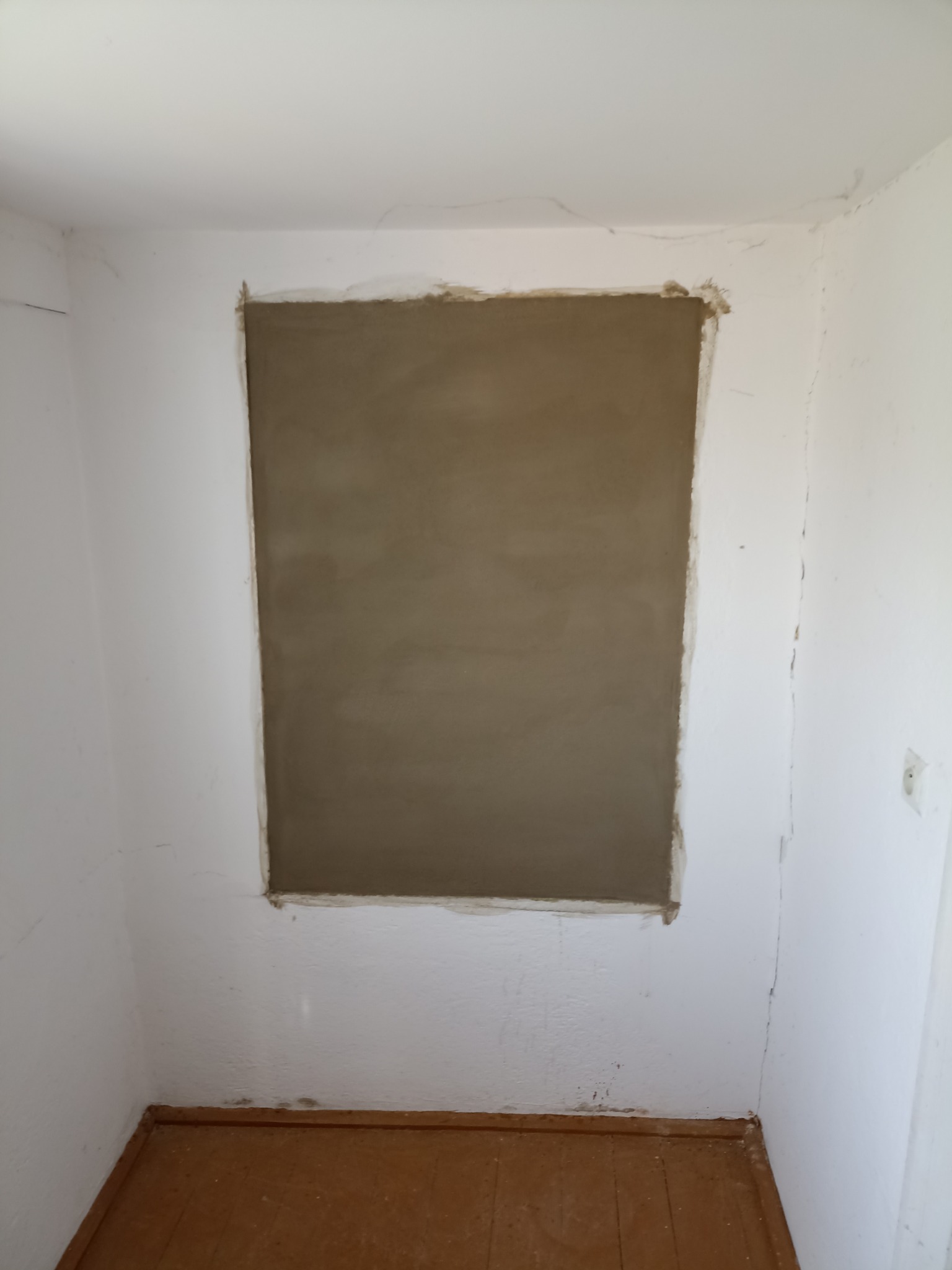 Świeżo nałożona, gładka warstwa szpachli w prostokątnym zagłębieniu ściany, przygotowana do dalszych prac wykończeniowych. Widoczne niedoskonałości starej farby.