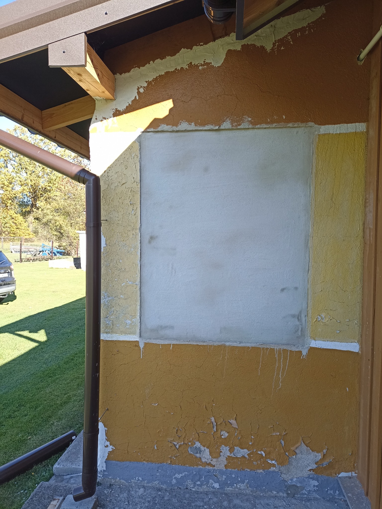 Fragment elewacji budynku w trakcie prac remontowych: otynkowane okno, łuszcząca się farba, brązowa rynna. Widoczne ślady wcześniejszych warstw farby.