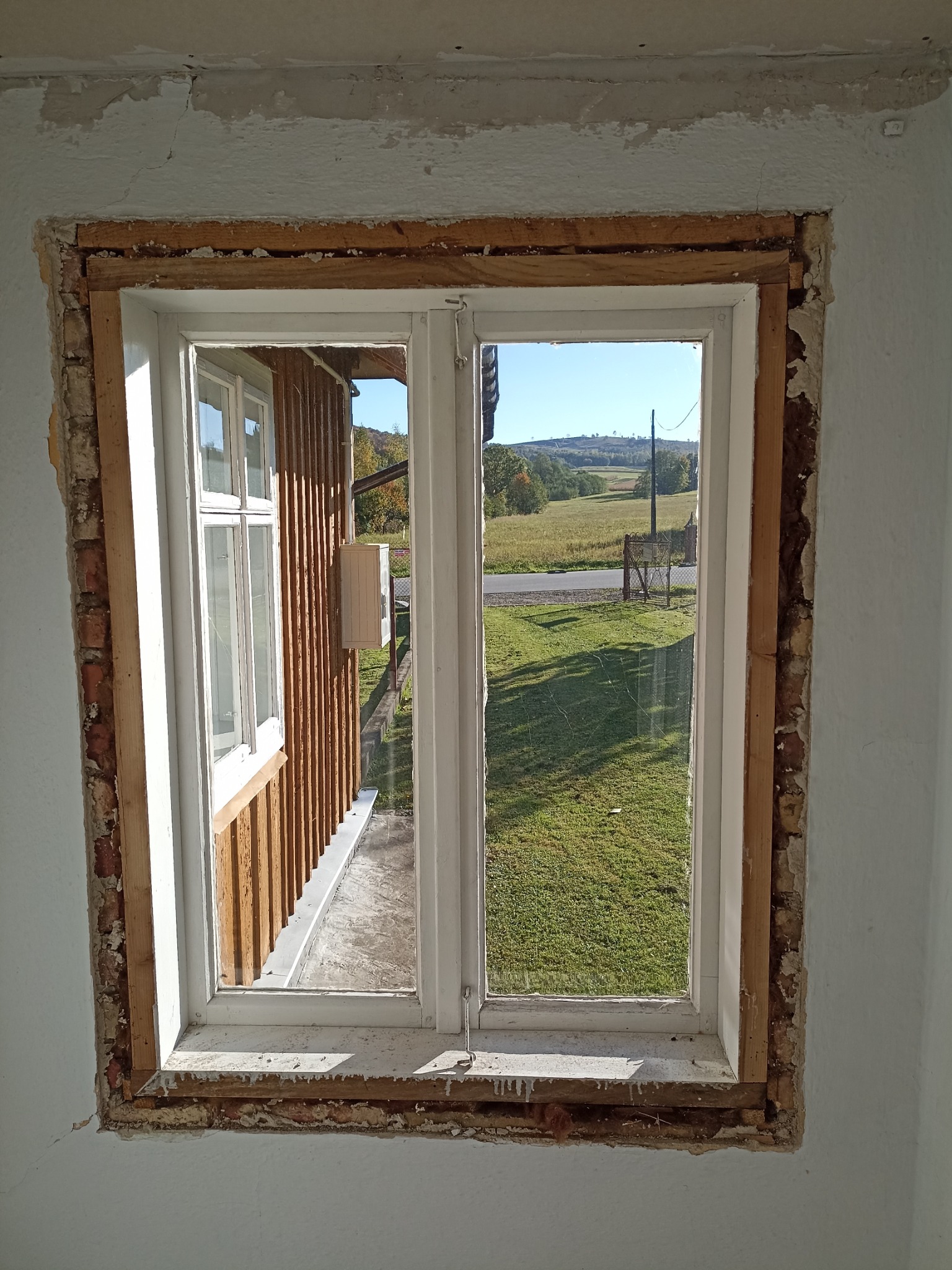 Białe okno z widokiem na zielony krajobraz, osadzone w surowym otworze okiennym z widocznymi śladami muru i resztkami zaprawy, ukazujące etap prac remontowych.