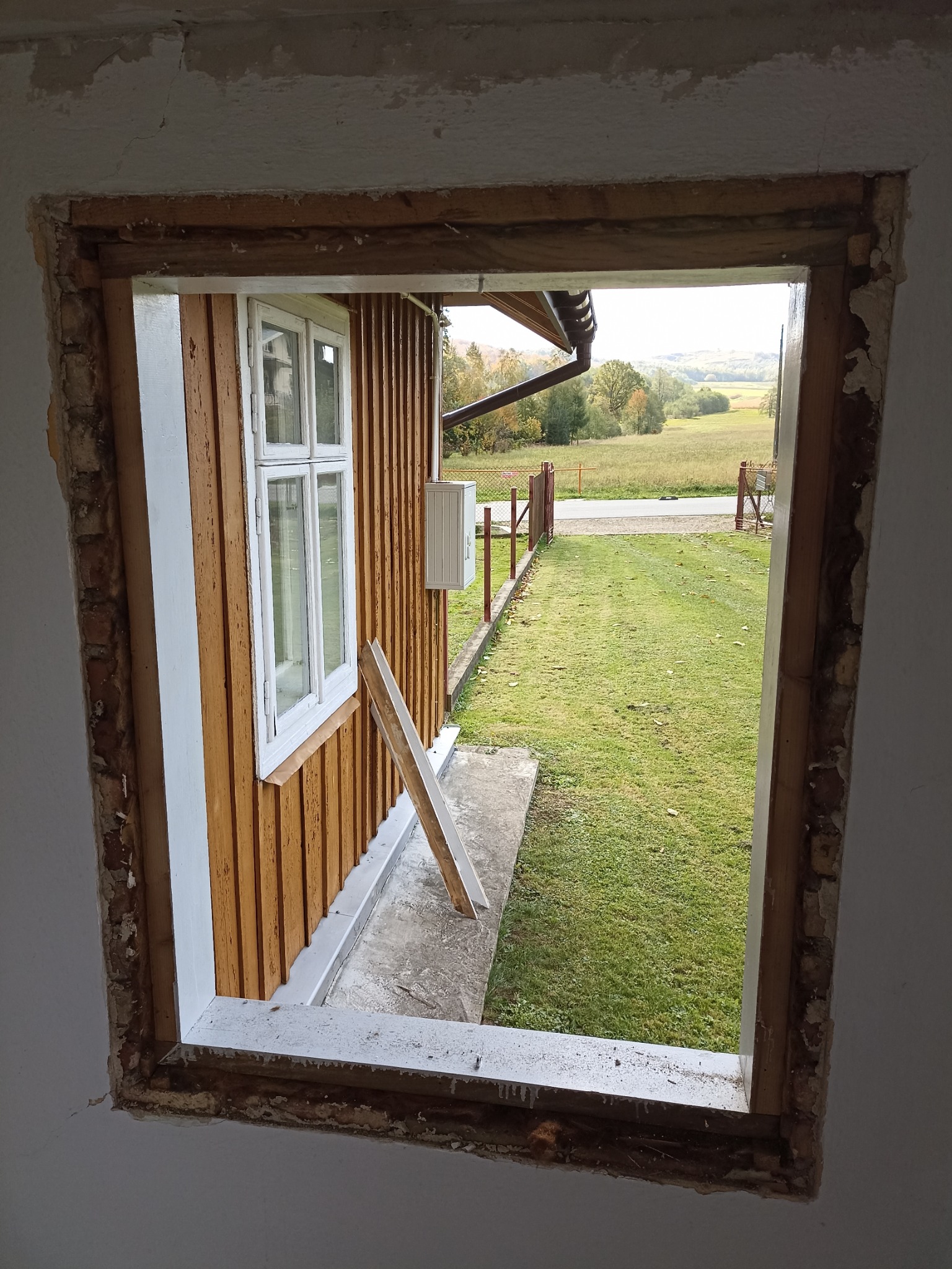 Otwór okienny w trakcie remontu, widoczne drewniane elementy konstrukcyjne i fragment elewacji z oknem na tle zielonego krajobrazu. Ślady po starym oknie.