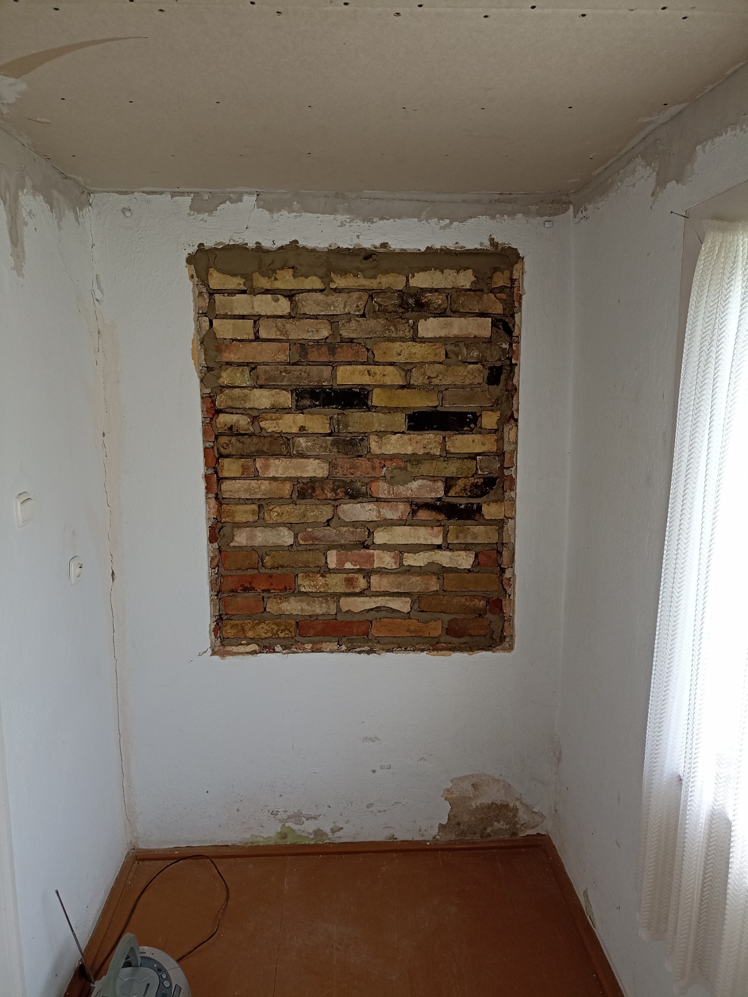 Fragment ściany z odsłoniętą cegłą w trakcie prac wykończeniowych. Widoczne ślady po usuniętych płytkach lub tynku, surowy stan wnętrza, przygotowanie do dalszych prac.