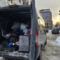 TwojeGraty - Szary bus z otwartymi drzwiami, wypełniony po brzegi różnymi przedmiotami, w tle budynki mieszkalne i śnieg. Widok z tyłu pojazdu.