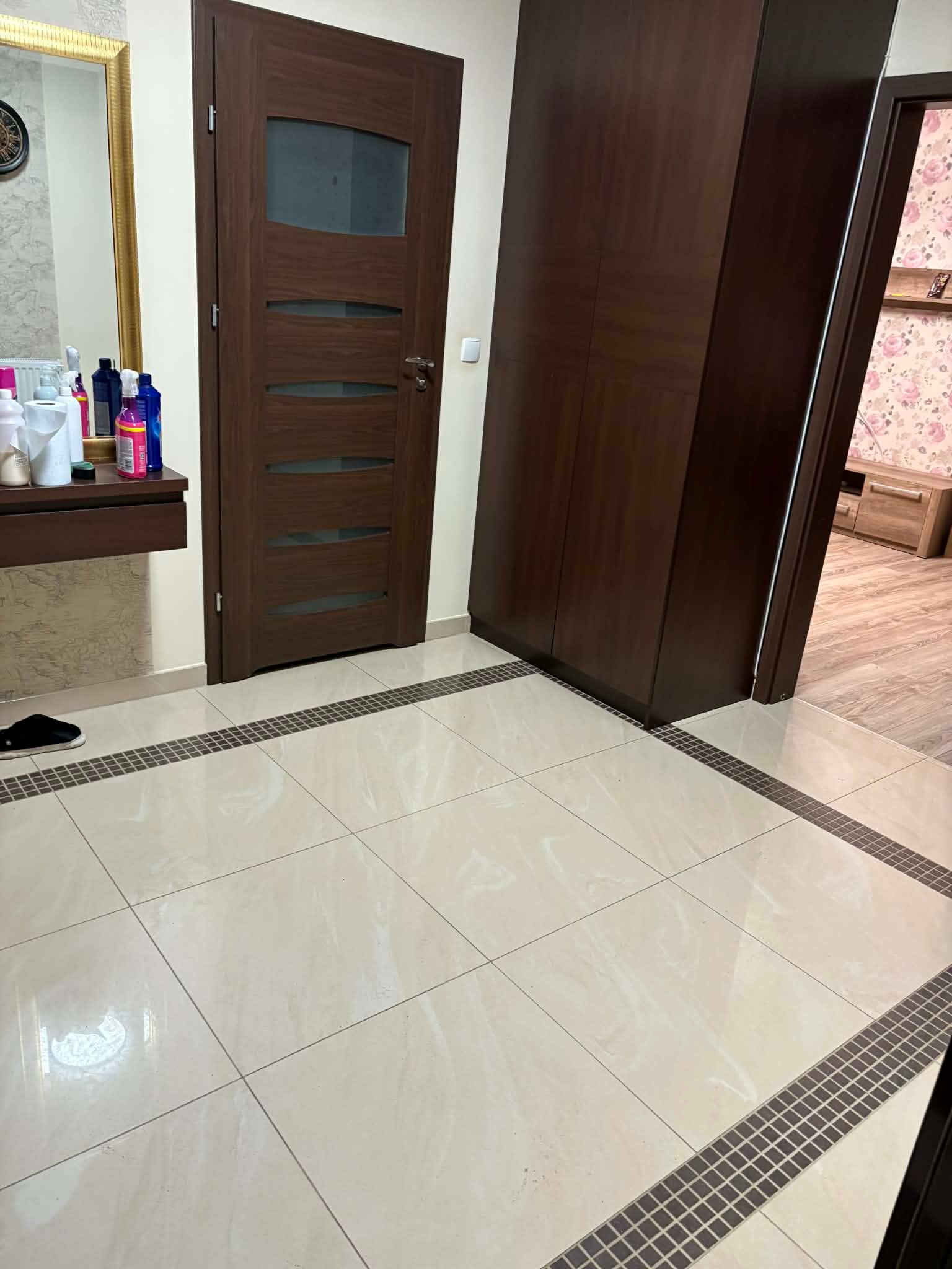 Ukończone wnętrze: beżowe płytki podłogowe z dekoracyjnym obramowaniem, brązowe drzwi z szybą i szafa wnękowa. Widoczny fragment toaletki z kosmetykami.