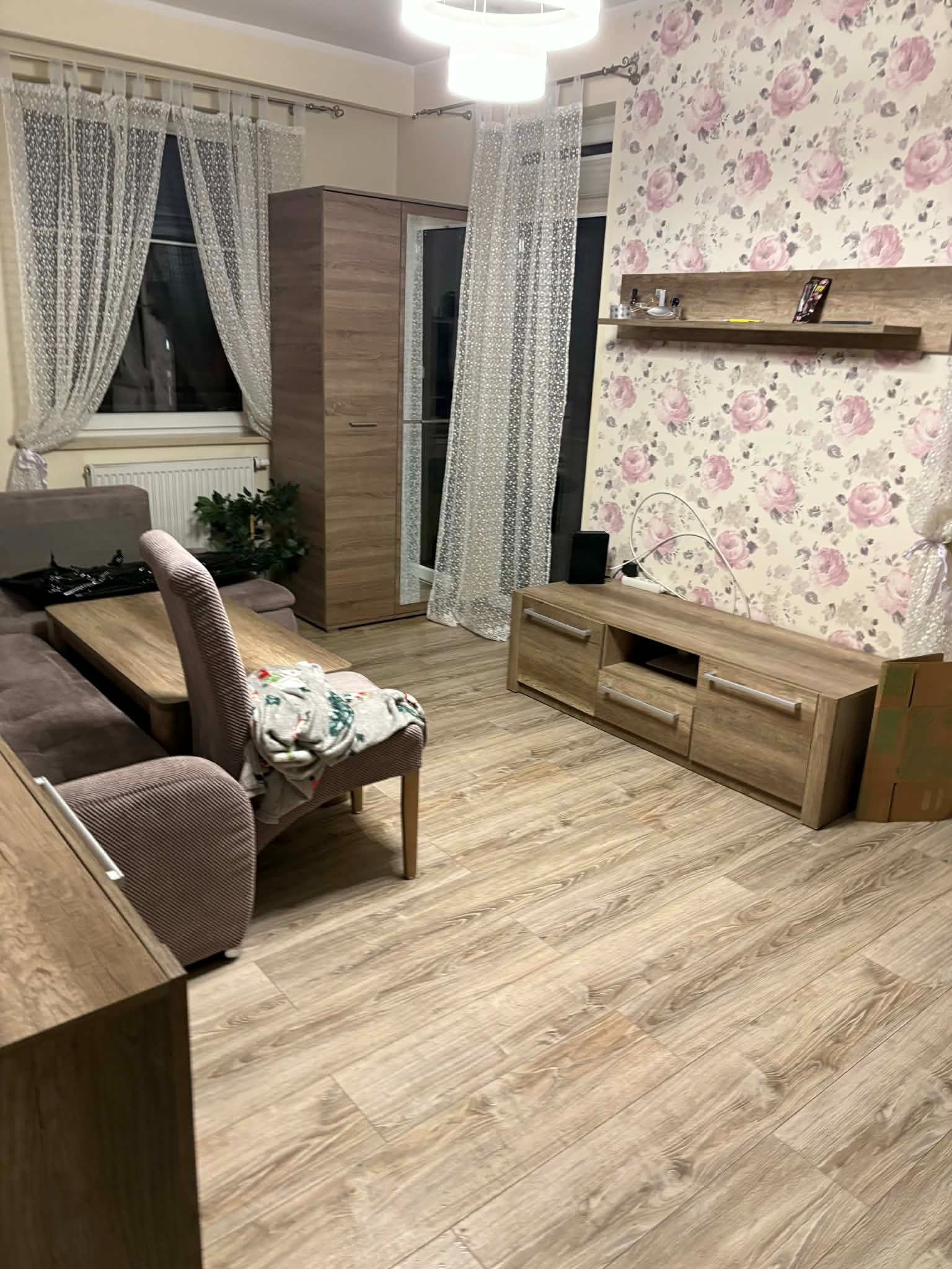 Umeblowane wnętrze pokoju dziennego z brązowymi meblami, wzorzystą tapetą w kwiaty i drewnianą podłogą. Widoczne okno z firankami i lampa sufitowa.