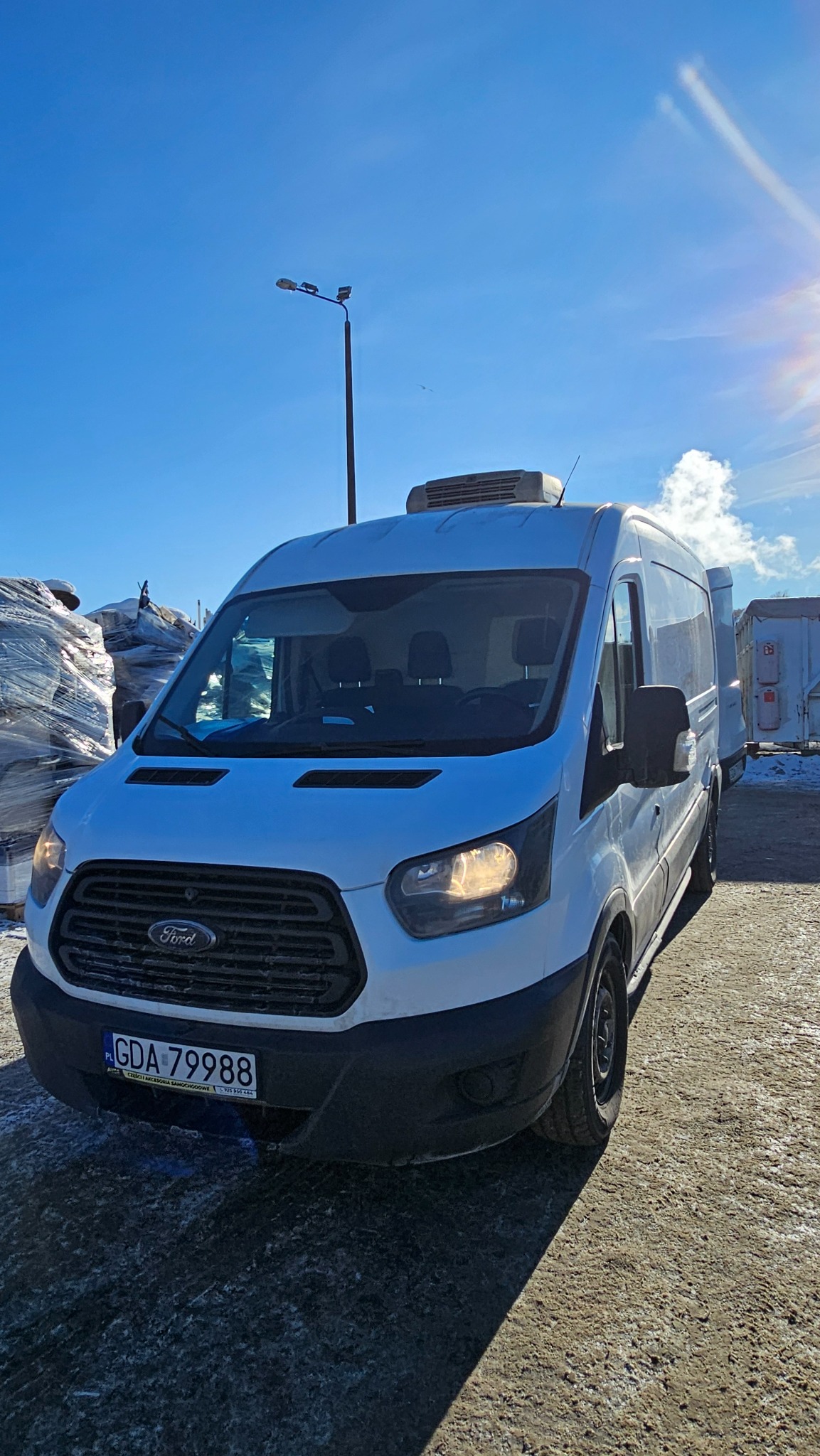 Biały furgon Ford Transit z klimatyzacją na dachu, zaparkowany na placu w słoneczny dzień. Widoczna tablica rejestracyjna GDA-79988 i fragmenty zapakowanych przedmiotów w tle.