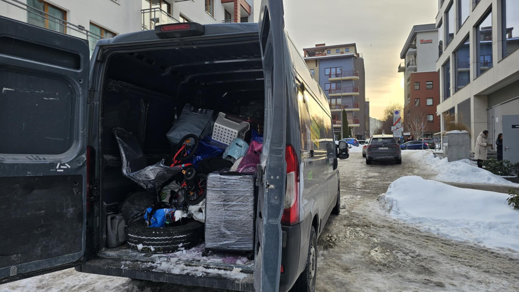 Szary bus z otwartymi drzwiami, wypełniony po brzegi różnymi przedmiotami, w tle budynki mieszkalne i śnieg. Widok z tyłu pojazdu.