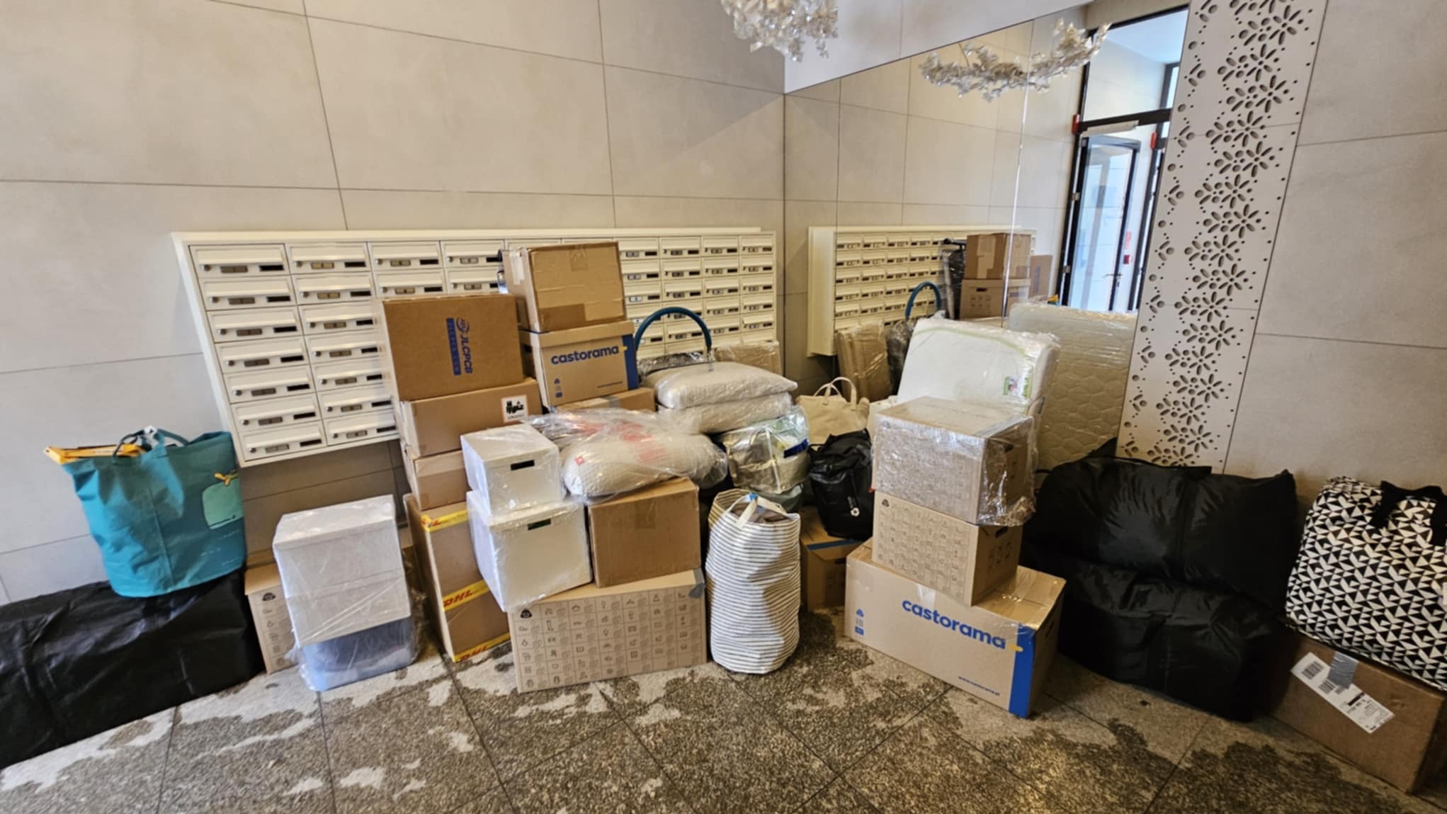 Stos kartonów i toreb w holu bloku, przygotowanych do transportu. Widoczne skrzynki pocztowe i drzwi wejściowe. Kartony Castorama.
