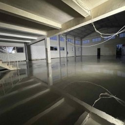 Karol Ślepecki - Wnętrze hali z nową, błyszczącą, szarą posadzką. Widoczne okna, filary i schody. Powierzchnia posadzki odbija światło, tworząc efekt lustra. Kable zwisają z sufitu.