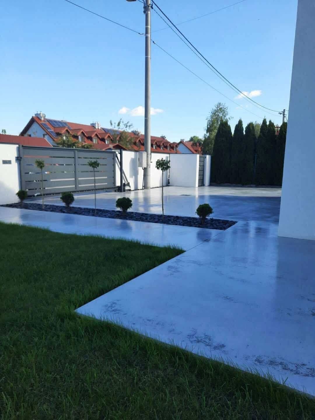 Gładka, szara posadzka przemysłowa na zewnątrz budynku, z elementami dekoracyjnymi z kamienia i zieleni. Nowoczesny design, brama w tle.