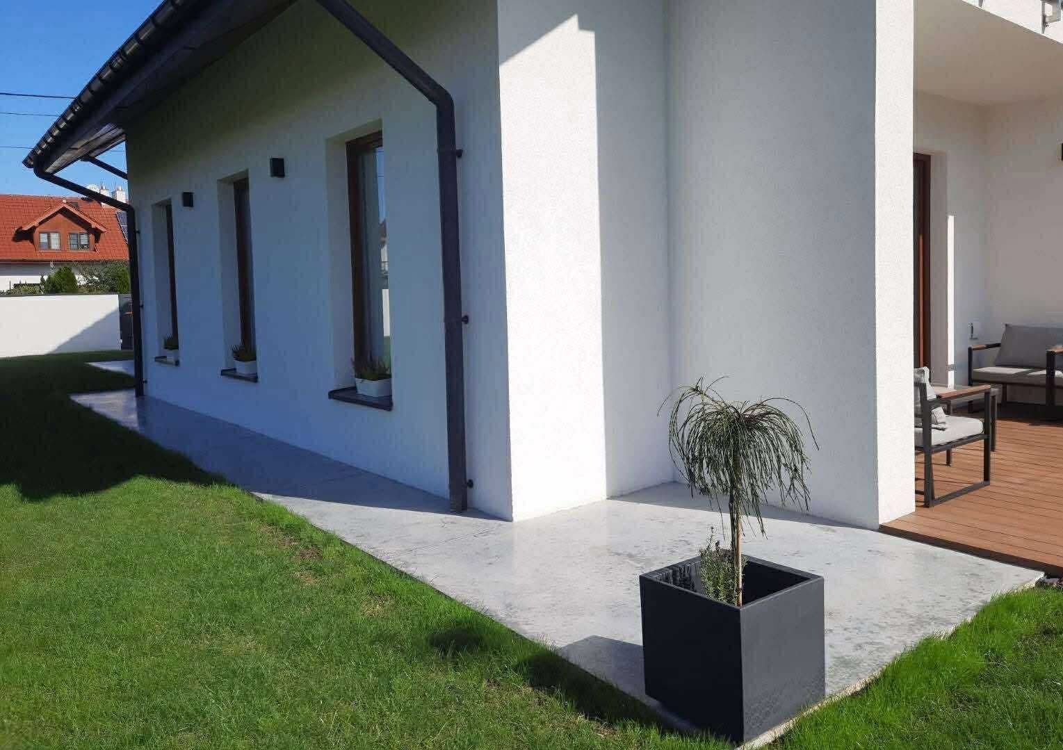 Jasnoszara, gładka posadzka na zewnątrz budynku z białymi ścianami i brązowymi oknami. W rogu donica z rośliną. Widoczny fragment trawnika i tarasu.