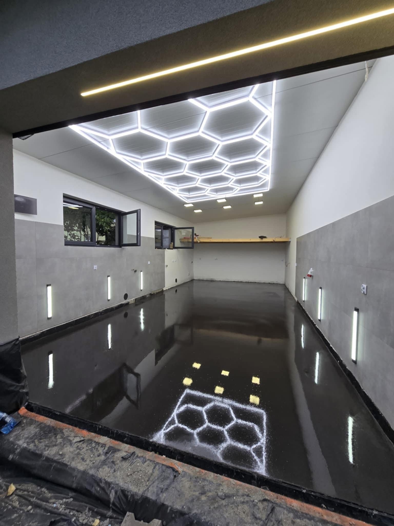 Błyszcząca, czarna posadzka przemysłowa w hali z oświetleniem LED w kształcie plastra miodu. Widoczne okna i listwy oświetleniowe na ścianach.