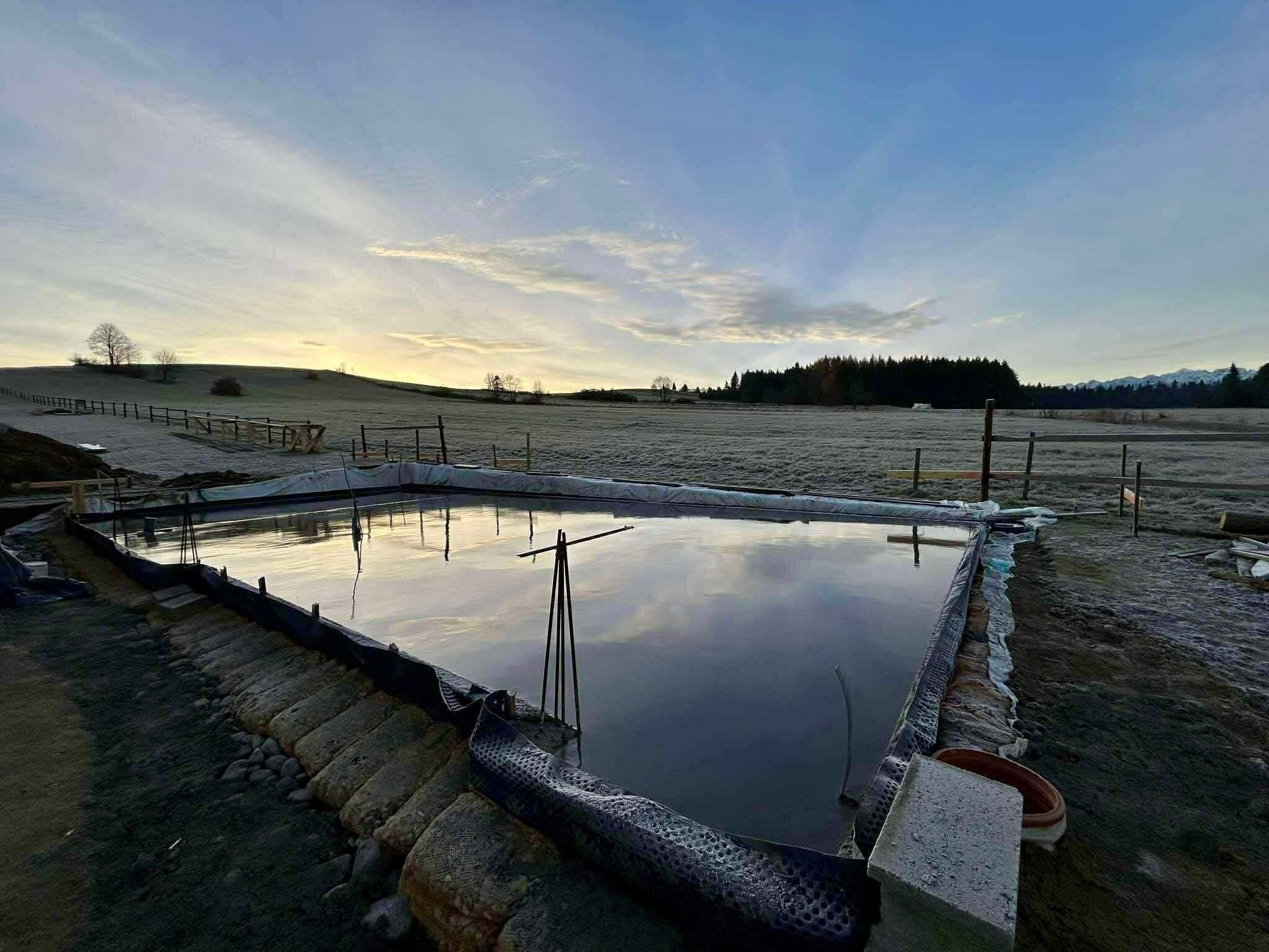 Wypełniony wodą, trójkątny zbiornik retencyjny w trakcie budowy, z widocznym zabezpieczeniem brzegów i odległym krajobrazem o wschodzie słońca.