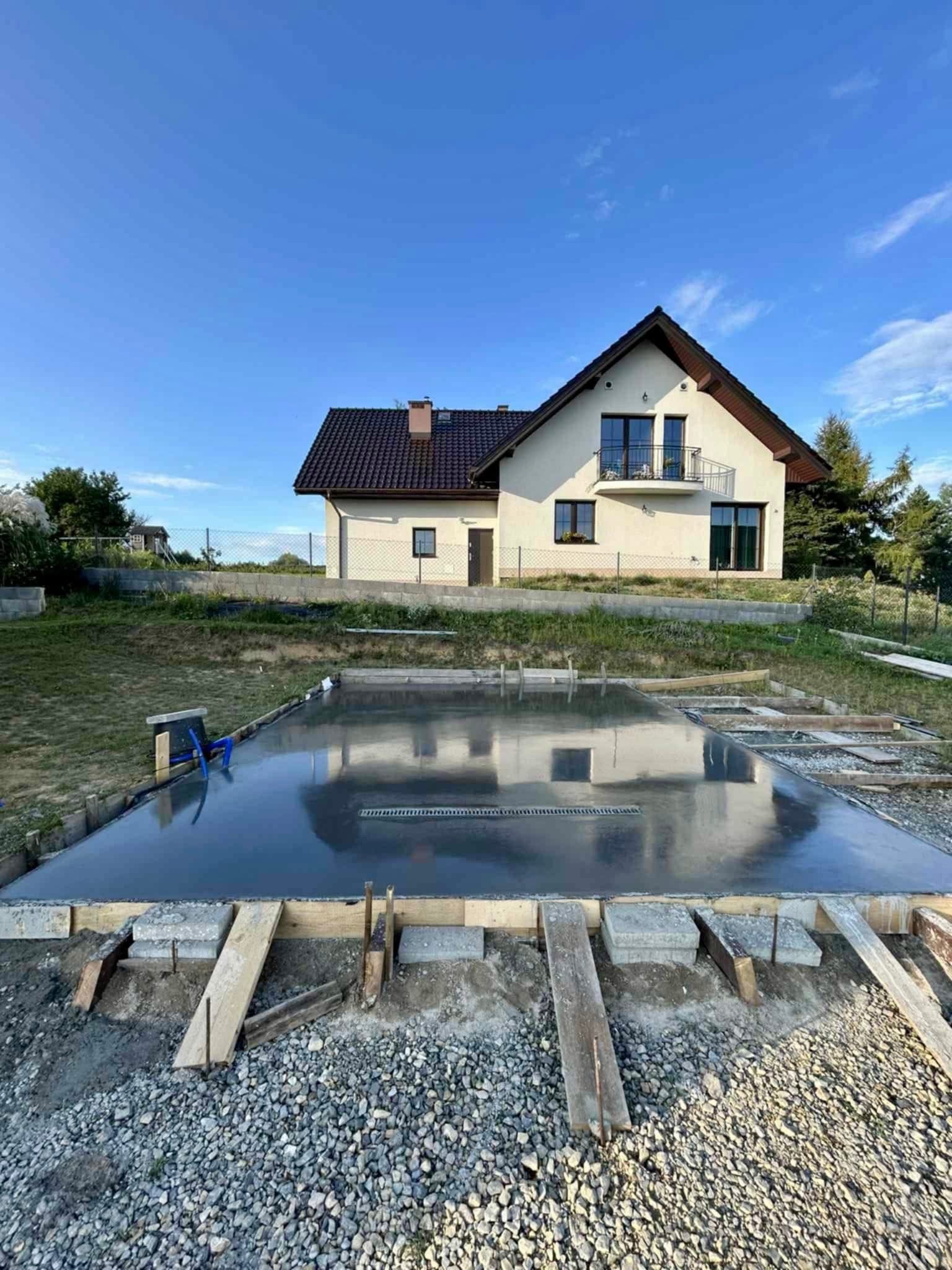 Świeżo wylana, gładka posadzka betonowa z odpływem liniowym na tle domu jednorodzinnego w słoneczny dzień. Widoczne szalunki i kamieniste podłoże.