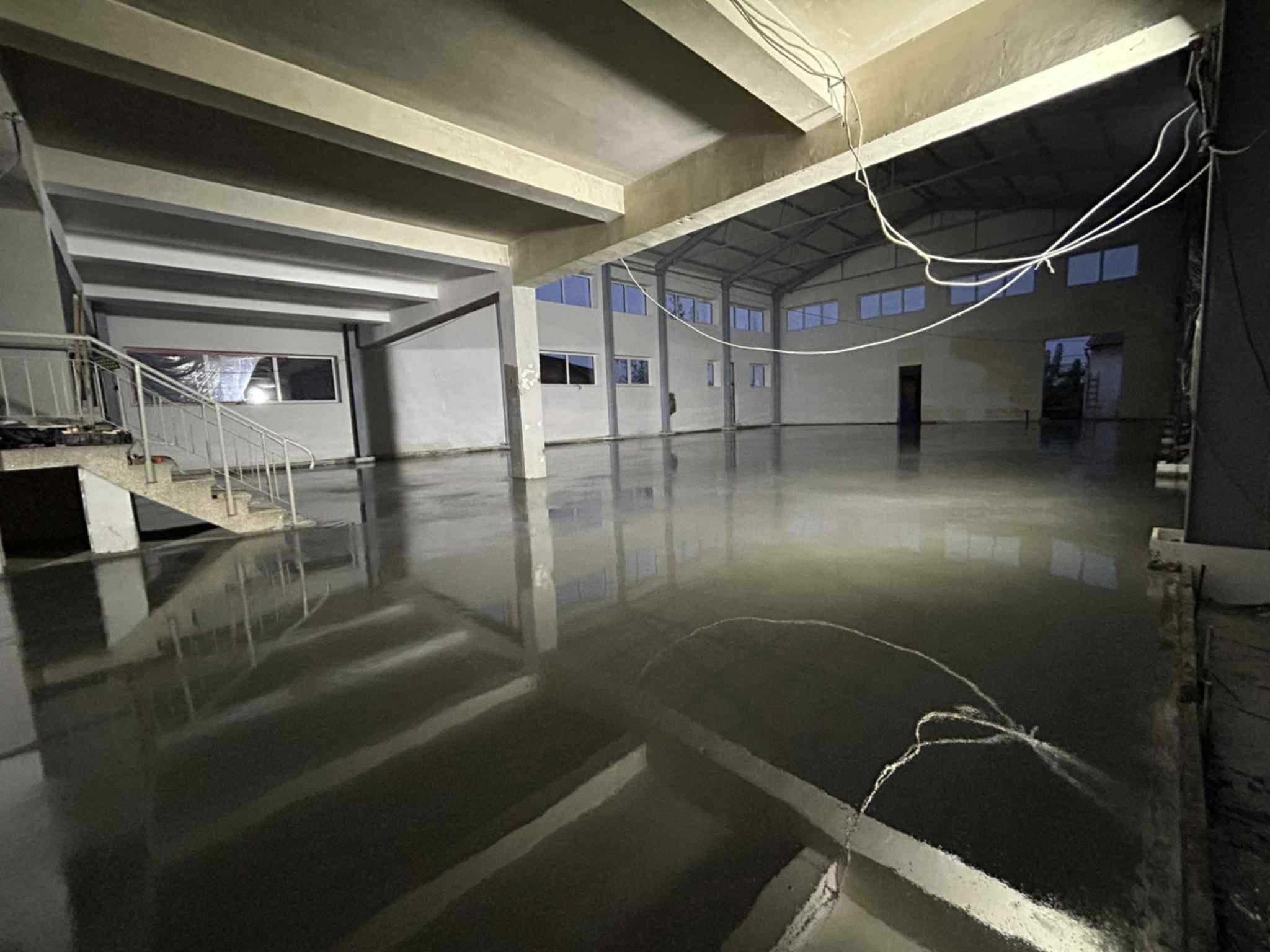 Wnętrze hali z nową, błyszczącą, szarą posadzką. Widoczne okna, filary i schody. Powierzchnia posadzki odbija światło, tworząc efekt lustra. Kable zwisają z sufitu.