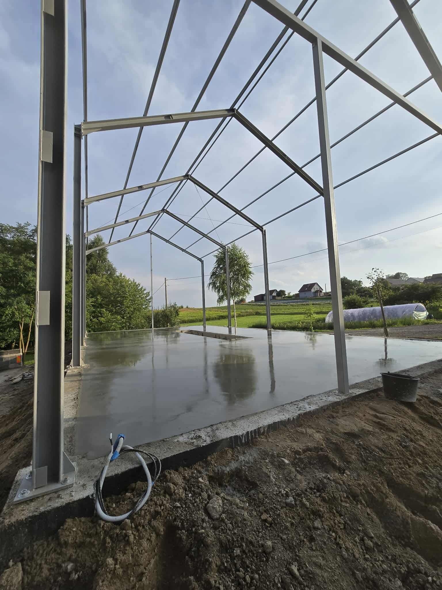 Świeżo wylana, gładka posadzka pod stalową konstrukcją hali, z widocznym kablem zasilającym i zielonym otoczeniem. Etap budowy hali.