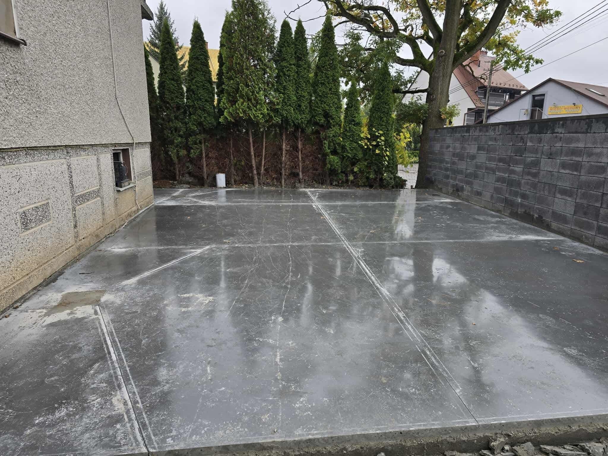 Szara, gładka posadzka przemysłowa podzielona na segmenty, z widocznymi refleksami i otoczeniem budynku oraz zielenią w tle. Widok z poziomu gruntu.