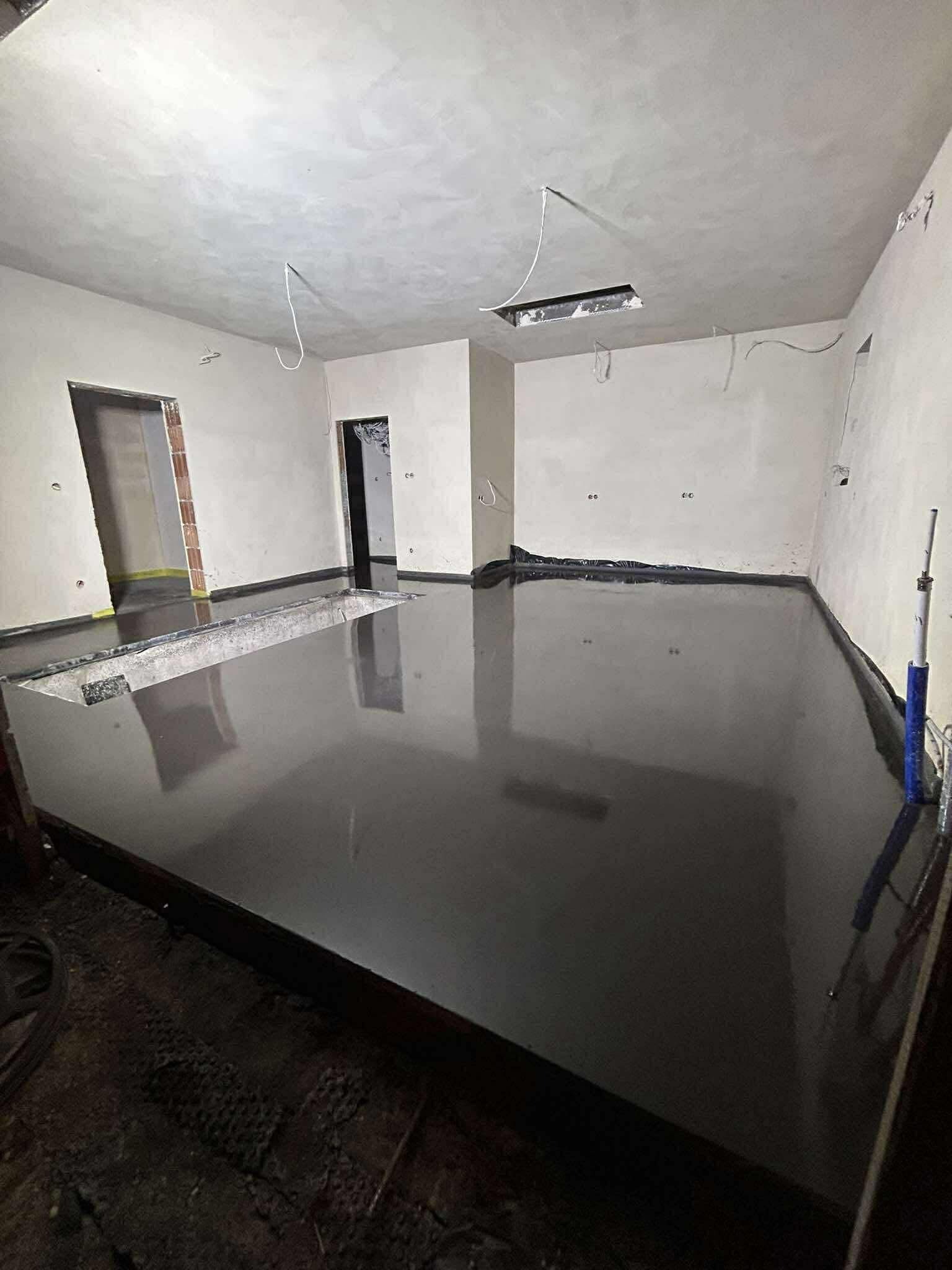 Świeżo wylana, gładka, ciemnoszara posadzka w surowym wnętrzu z widocznymi otworami w ścianach i suficie. Na podłodze widoczne narzędzia i folia ochronna.