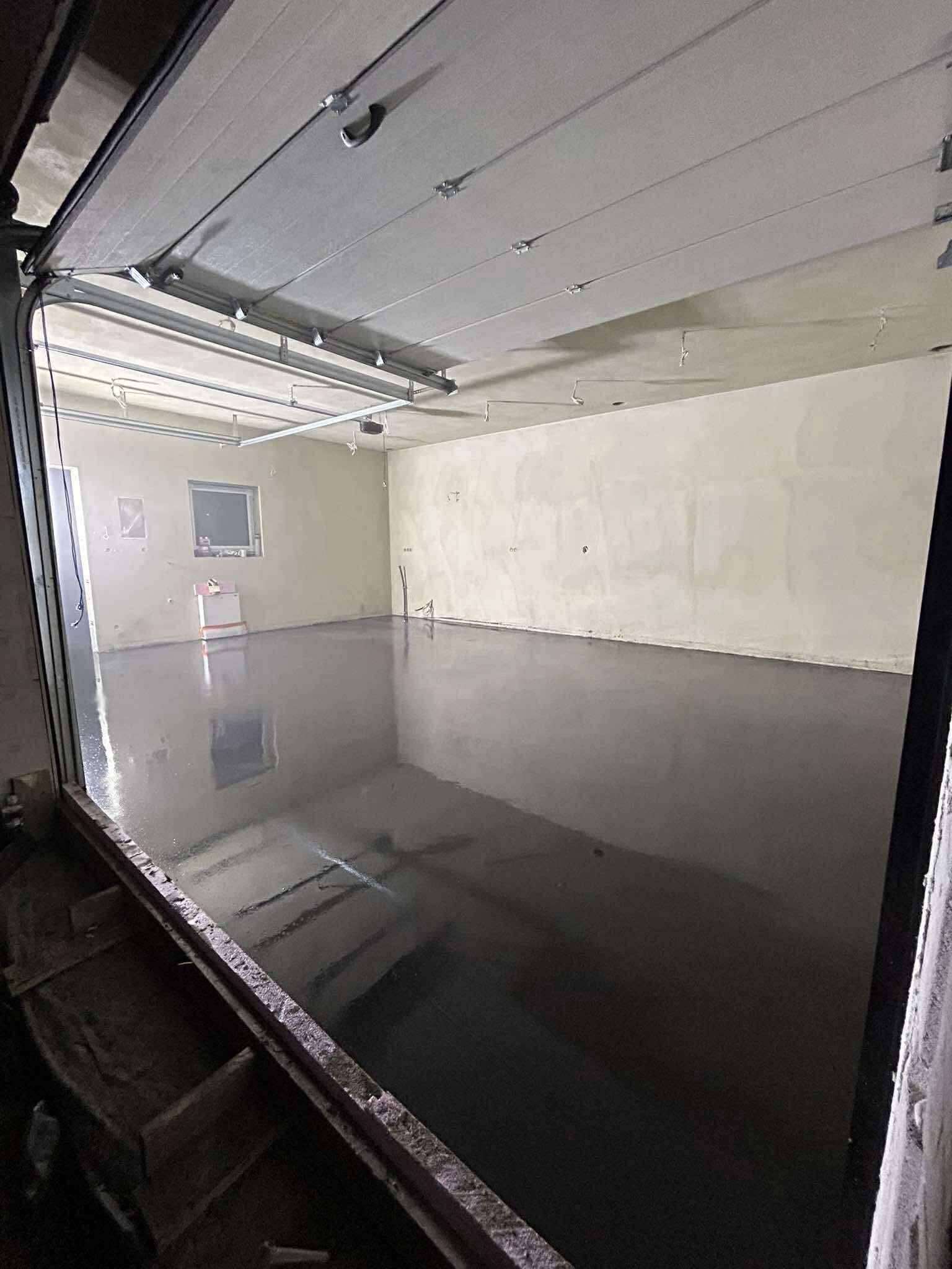 Błyszcząca, ciemnoszara posadzka w pustym pomieszczeniu z otwartą bramą garażową. Widoczne ślady po pracach wykończeniowych na ścianach.