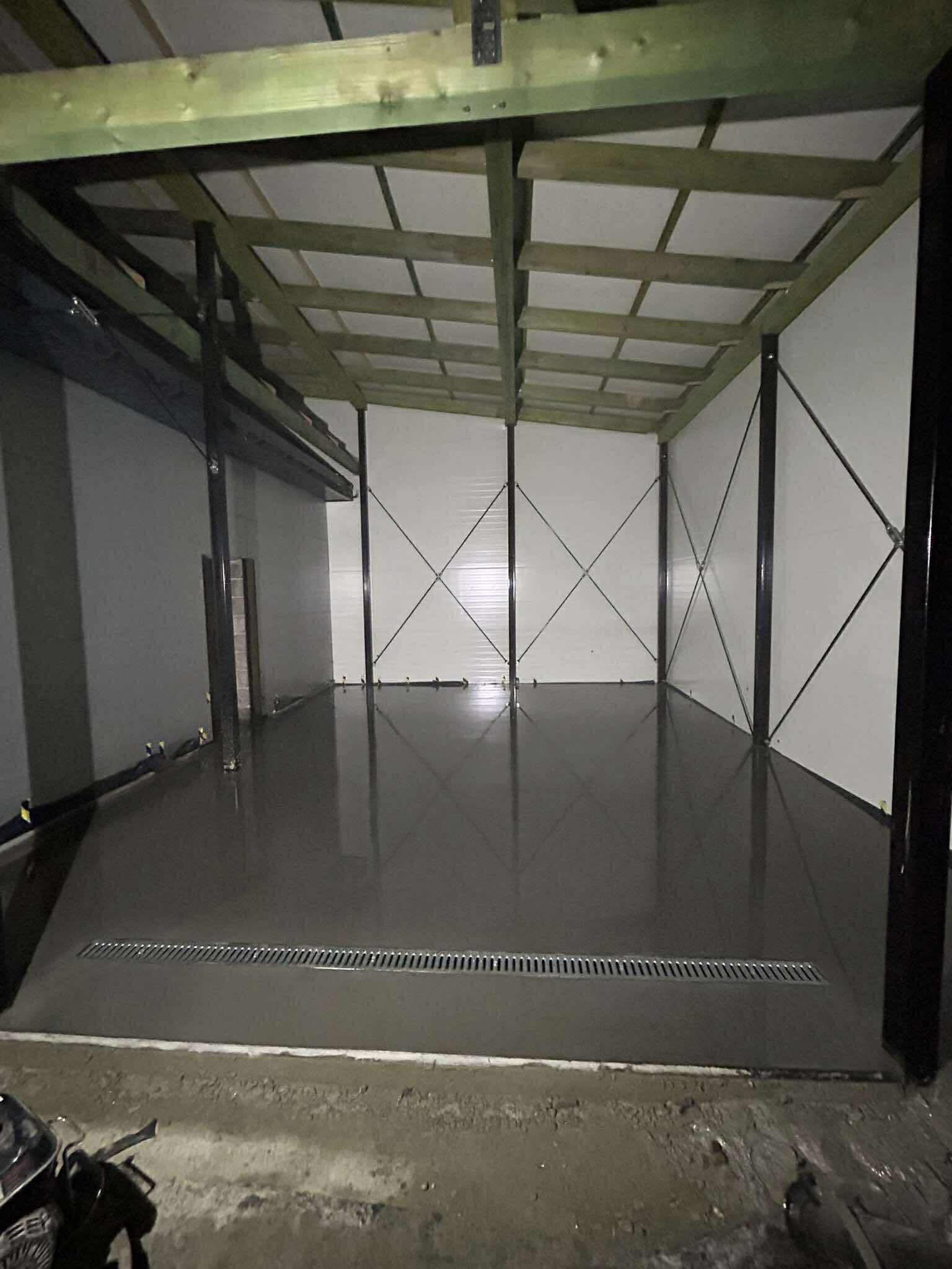 Świeżo wylana, gładka posadzka przemysłowa w hali z metalową konstrukcją i kratką odpływową. Wilgotna powierzchnia odbija światło, podkreślając równość wylewki.