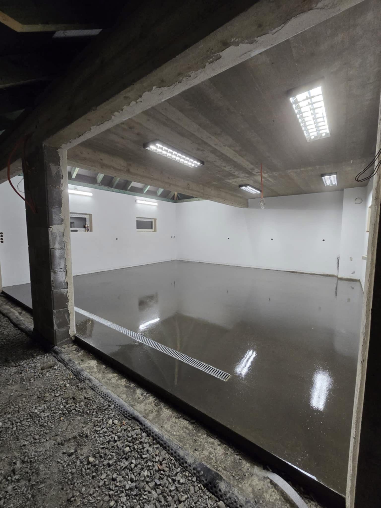 Świeżo wylana, błyszcząca posadzka w surowym wnętrzu z widocznym odpływem liniowym i betonowym stropem z oświetleniem. Widoczne niedokończone elementy konstrukcyjne.