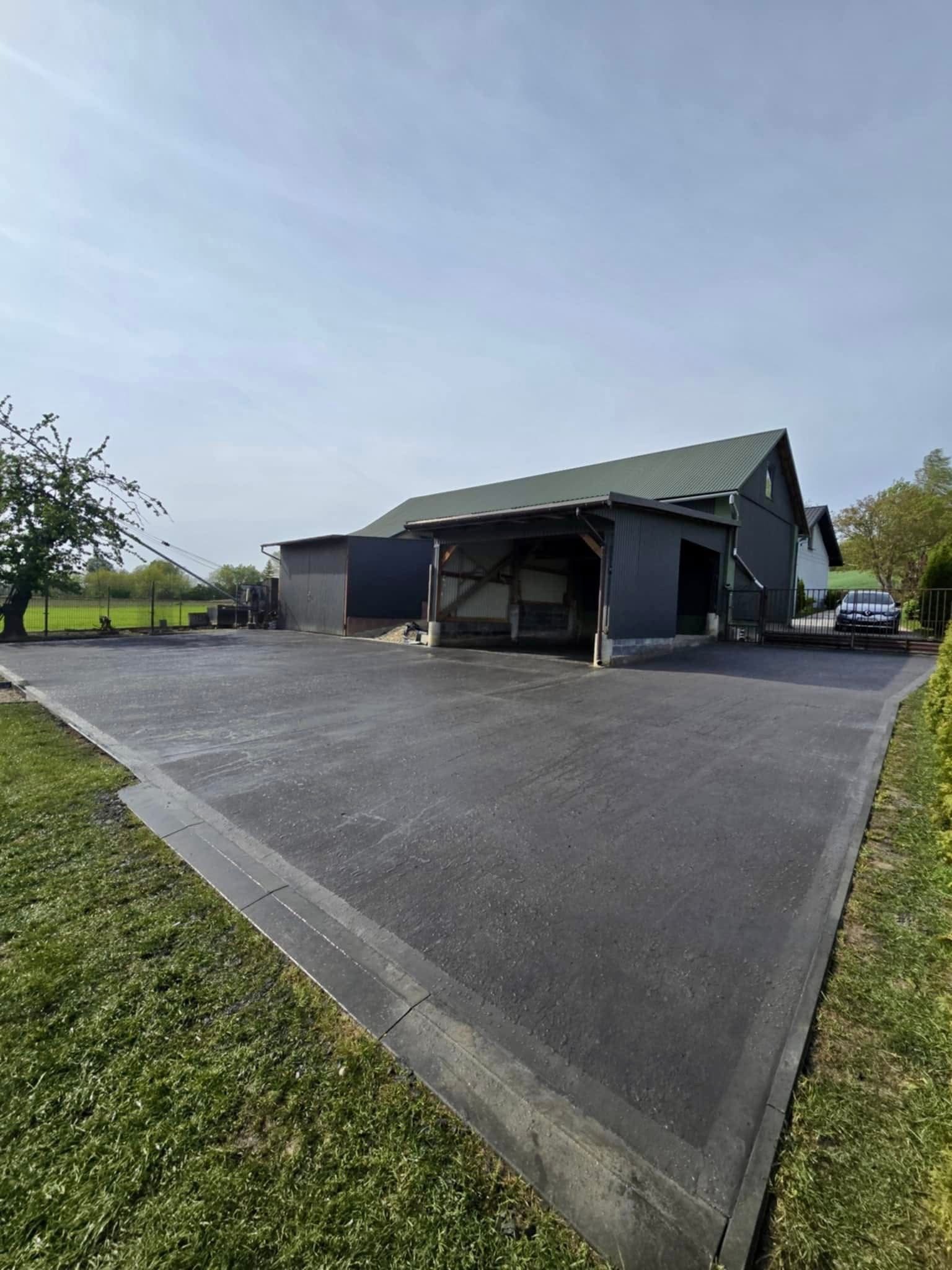 Gładka, ciemna posadzka przemysłowa na zewnątrz budynku gospodarczego z zielonym dachem, otoczona trawnikiem i drzewami w słoneczny dzień. Widoczny fragment ogrodzenia i samochód.