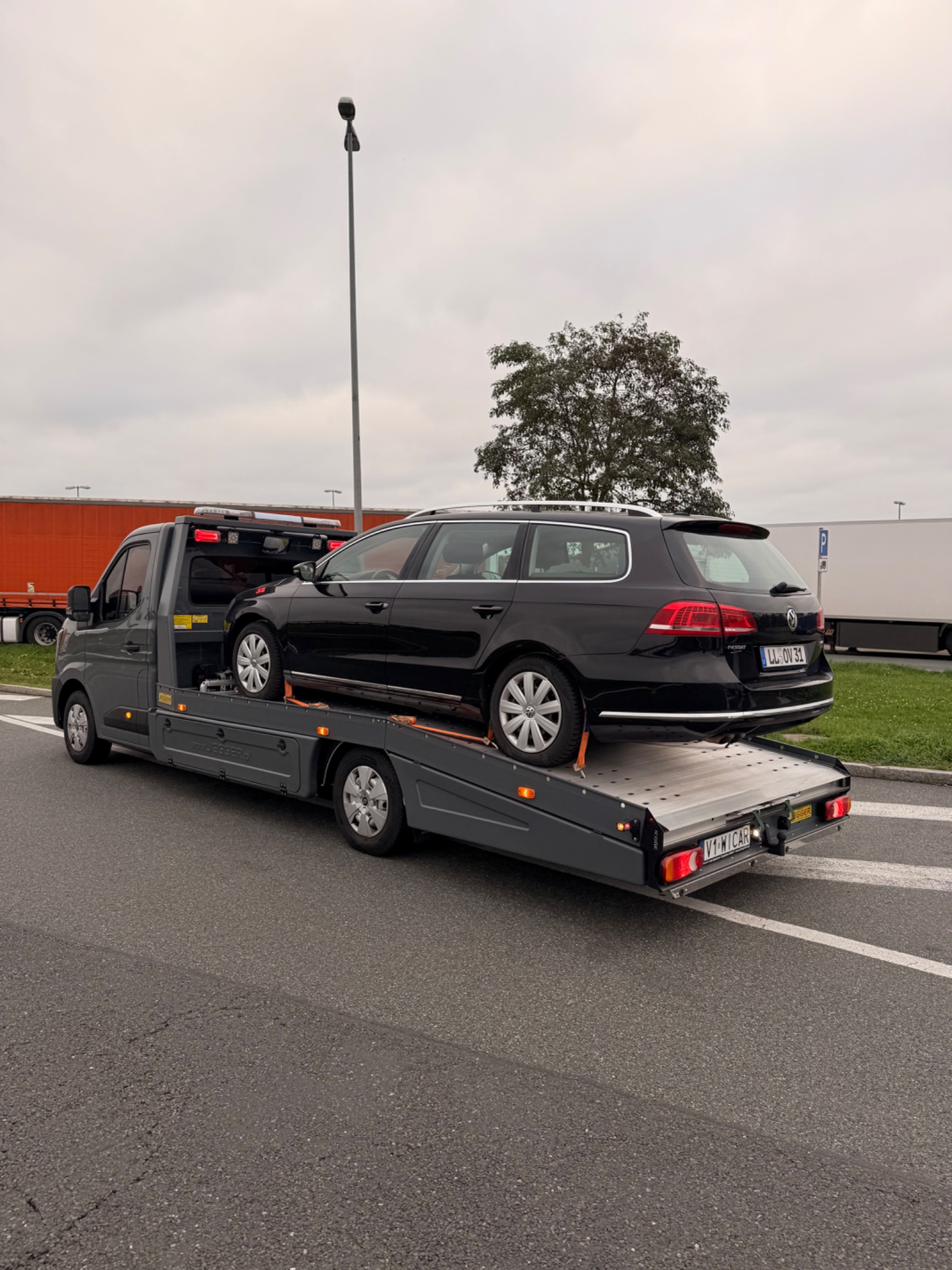 Czarna laweta z czarnym kombi Volkswagen Passat na platformie, widok z tyłu, tablica rejestracyjna LL OV 31, szare niebo w tle.