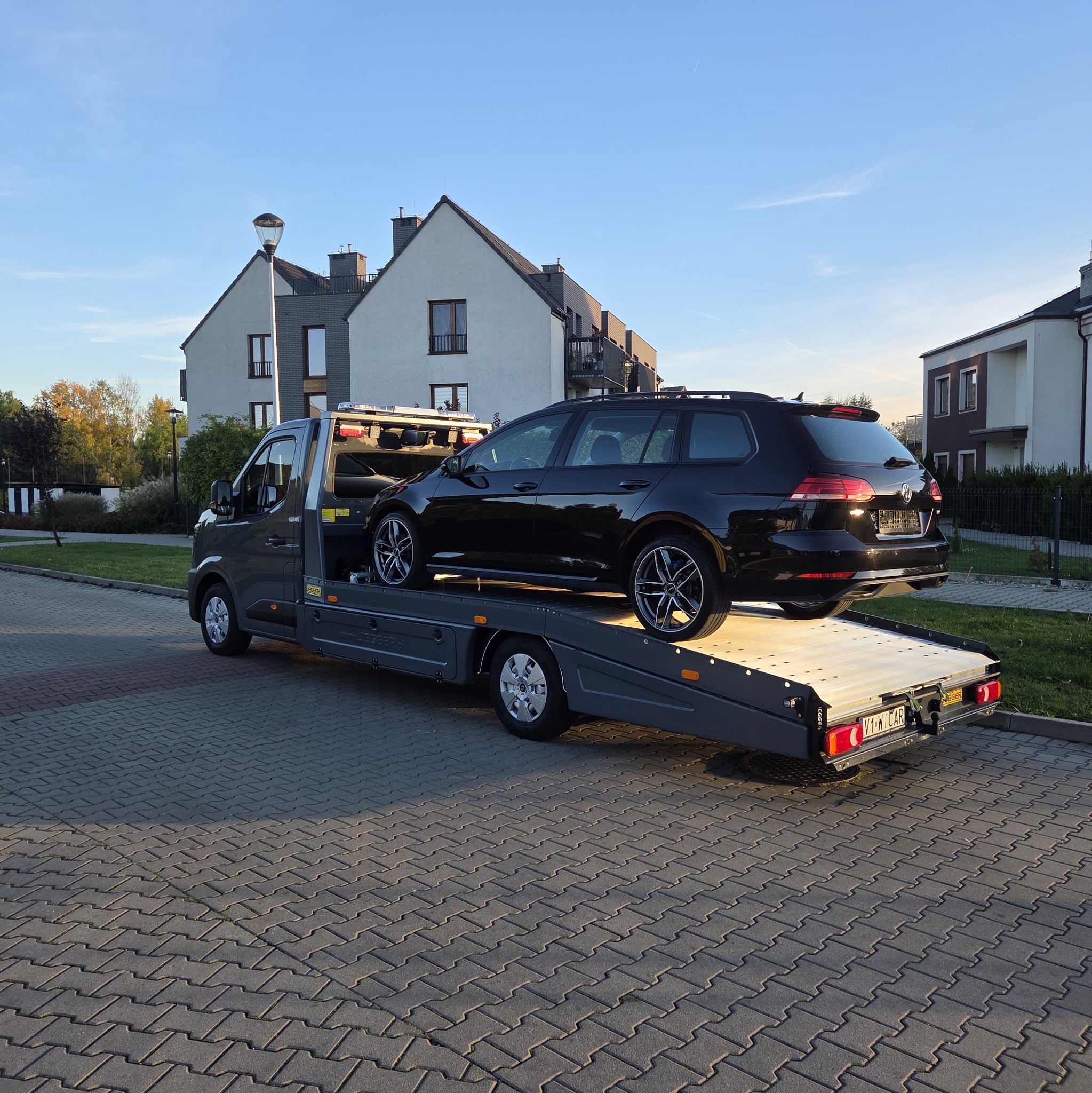 Czarny samochód kombi na szarej lawecie w otoczeniu nowoczesnych domów jednorodzinnych i kostki brukowej. Słoneczny dzień, błękitne niebo.