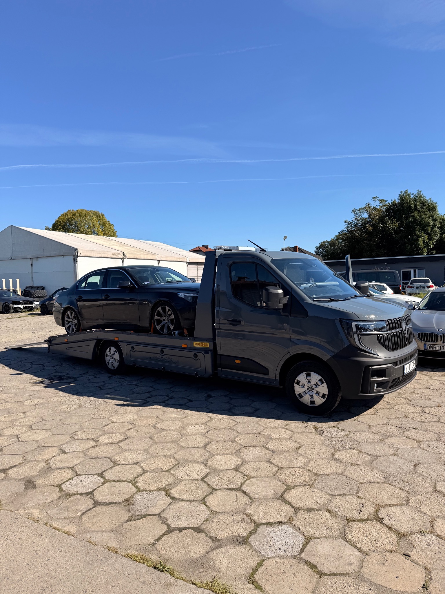 Szara laweta z czarnym samochodem osobowym na platformie, zaparkowana na placu z betonowej kostki, w tle budynki i drzewa, słoneczny dzień.