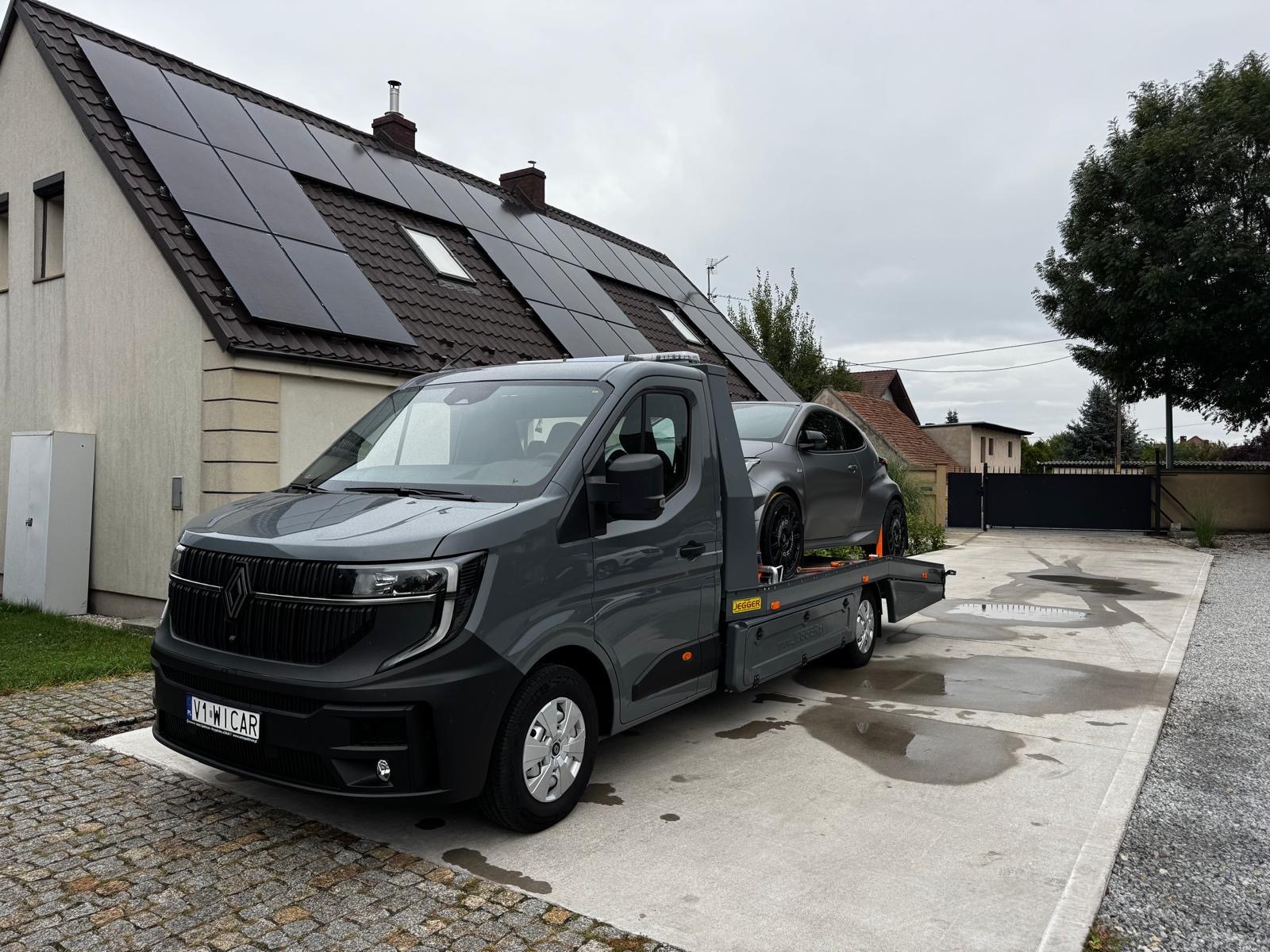 Szara laweta Renault z szarym sportowym autem na platformie, zaparkowana na betonowym podjeździe przed domem z panelami słonecznymi na dachu.